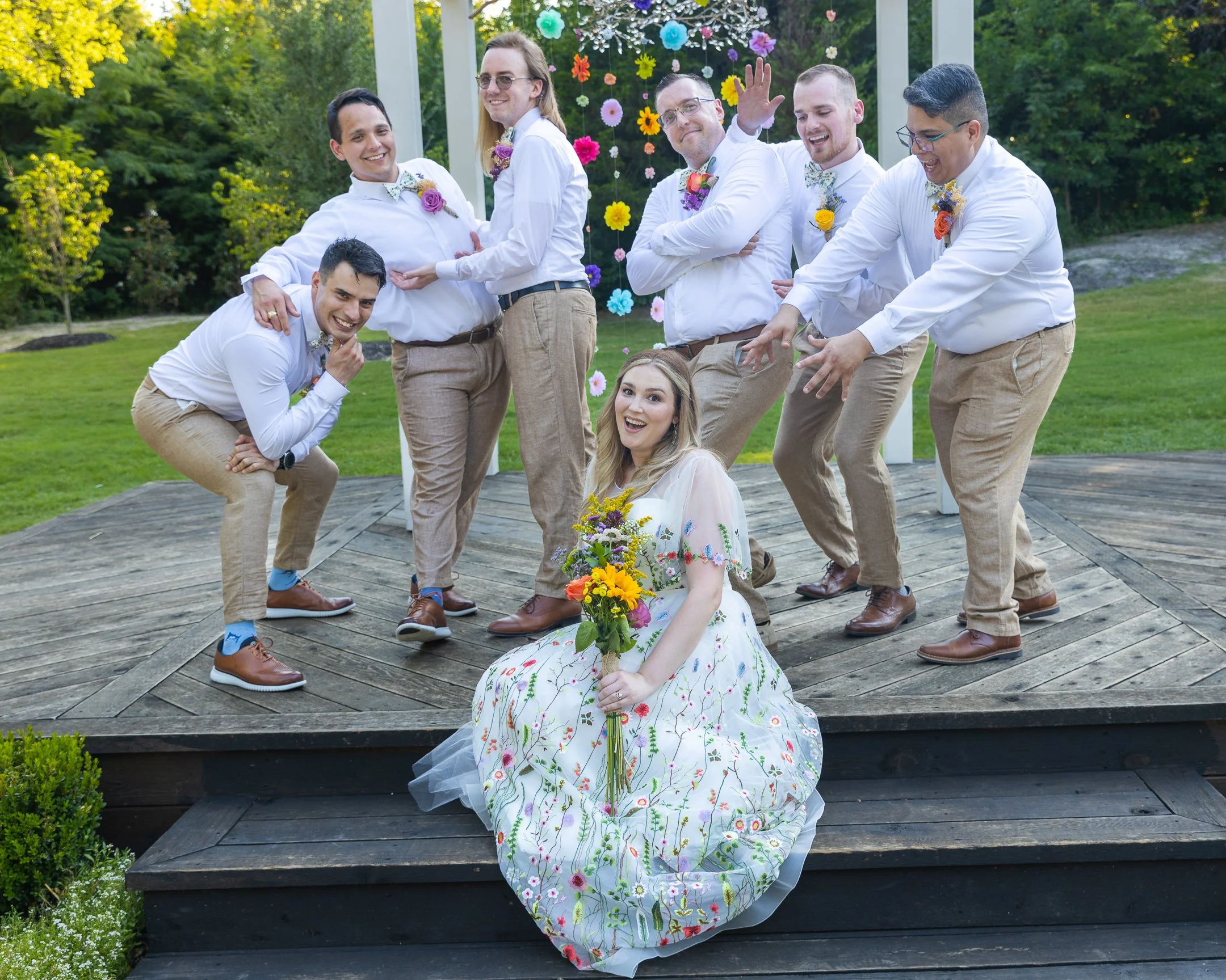 A bride with a bouquet sitting on the wooden steps in front of a group of groomsmen and bridesmaids, outdoors on a wooden stage with a colorful flower backdrop, all smiling and playful.