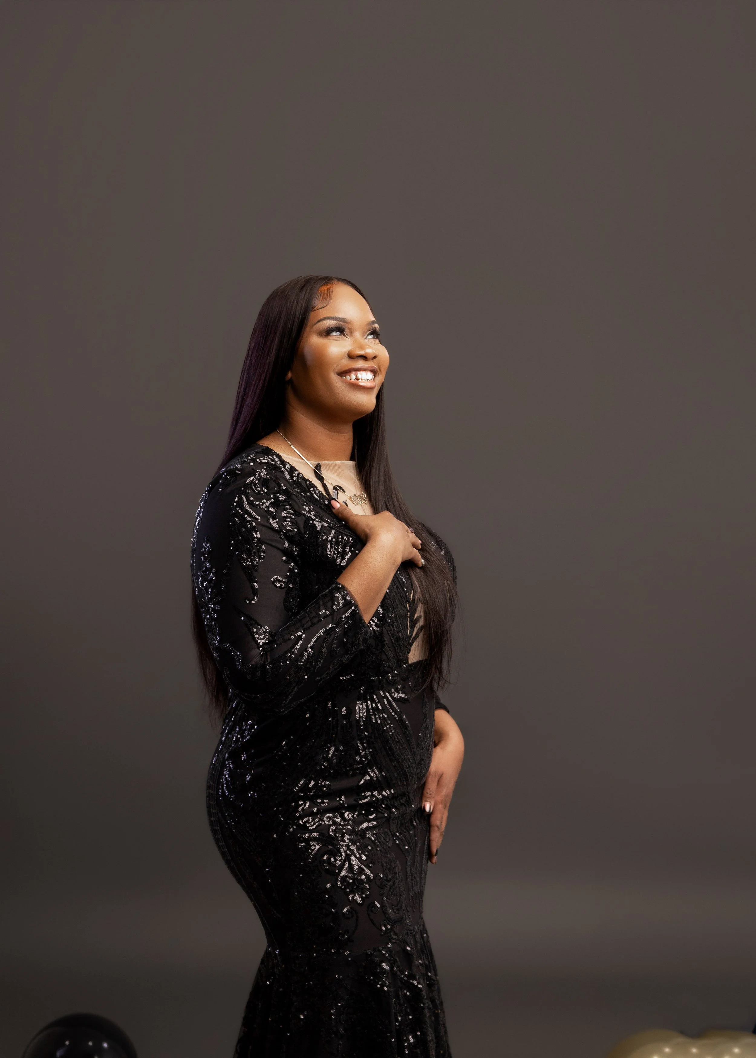 A woman dressed in a black, sequined gown with a beige top underneath, posing against a plain gray background, smiling with her hand over her chest.