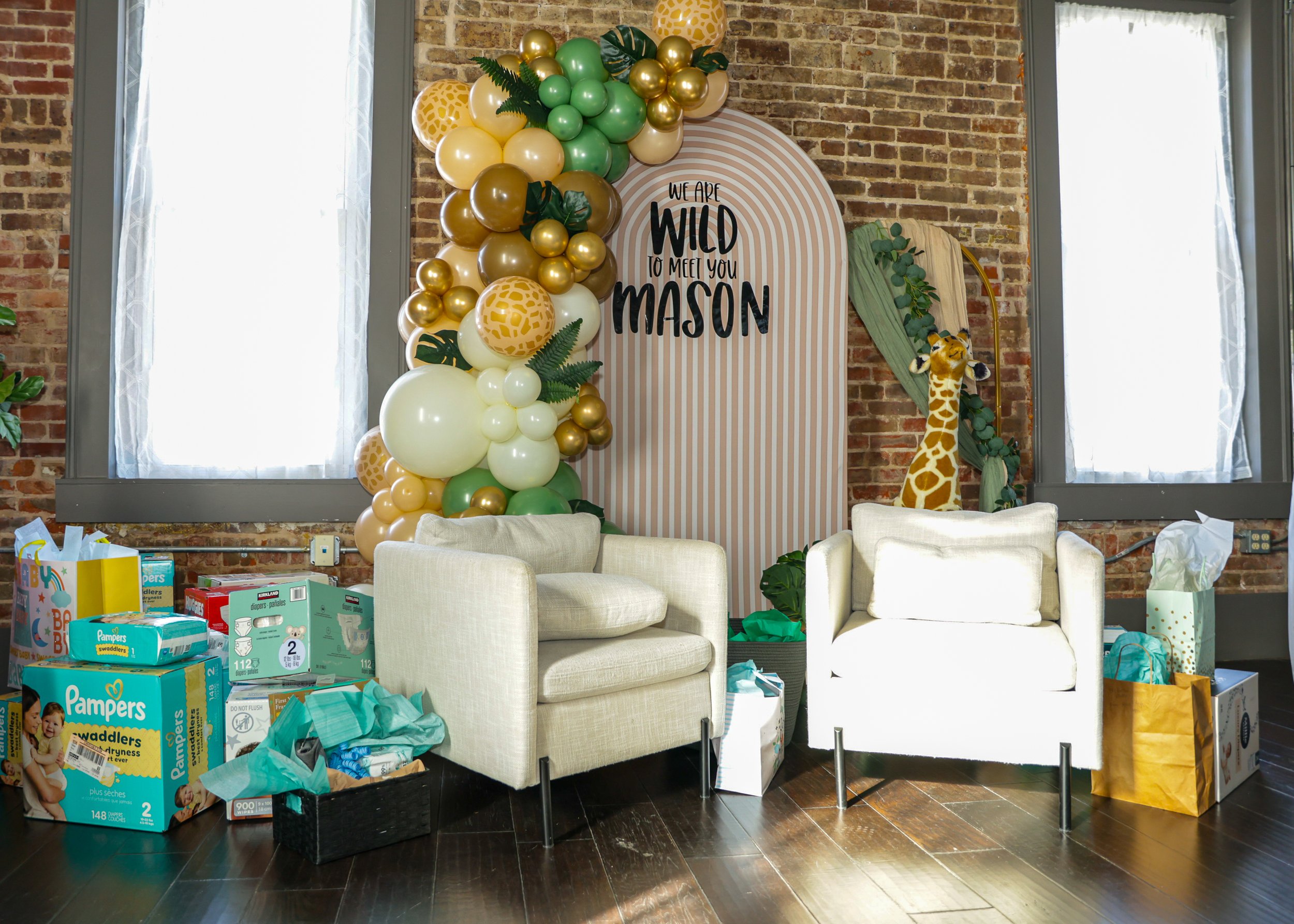 A decorated indoor space with a balloon arch featuring green, gold, and cream-colored balloons, large leaves, and a sign that reads "We are WILD to meet you Mason." There is a giraffe plush toy sitting on a green pillar on the right side, and various