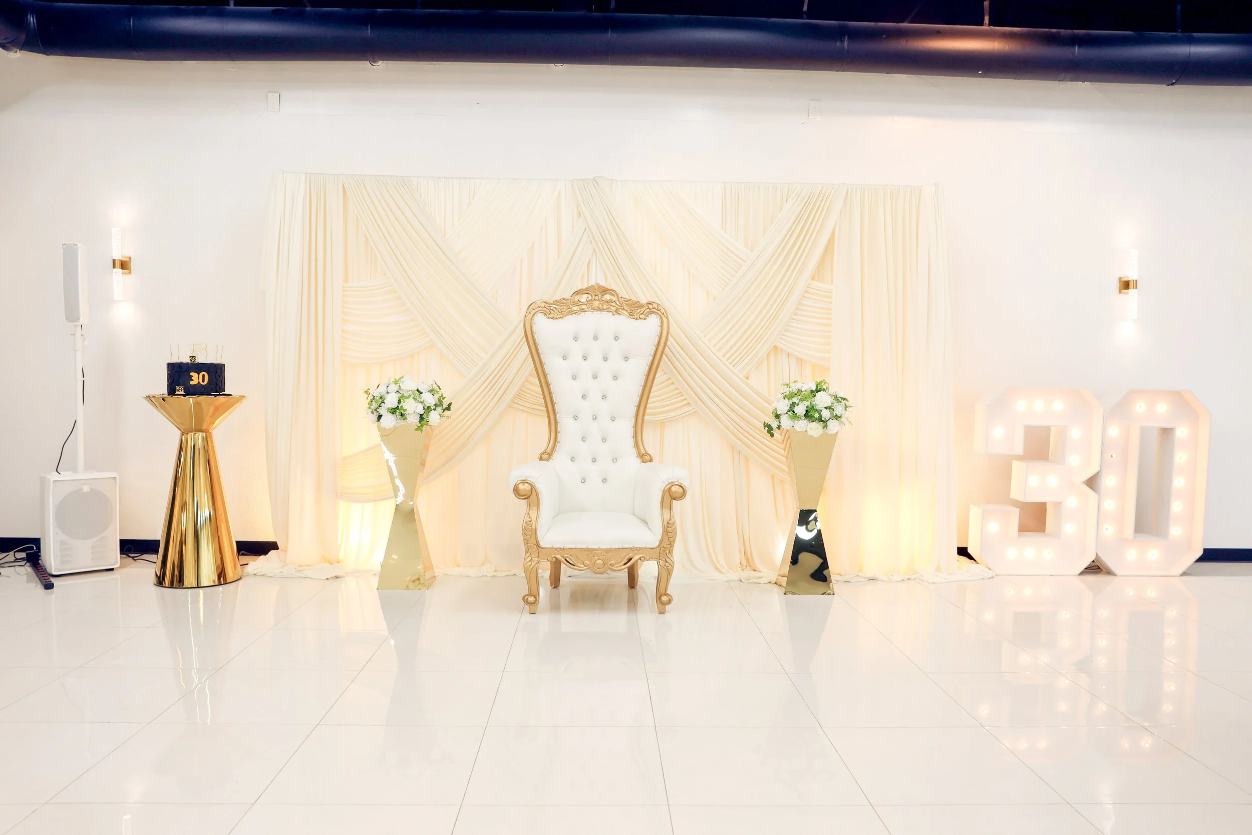 Elegant white high-backed armchair with gold trim positioned in front of a cream-colored draped curtain backdrop, decorated with flowers on either side, and illuminated marquee letters spelling out the number 30.