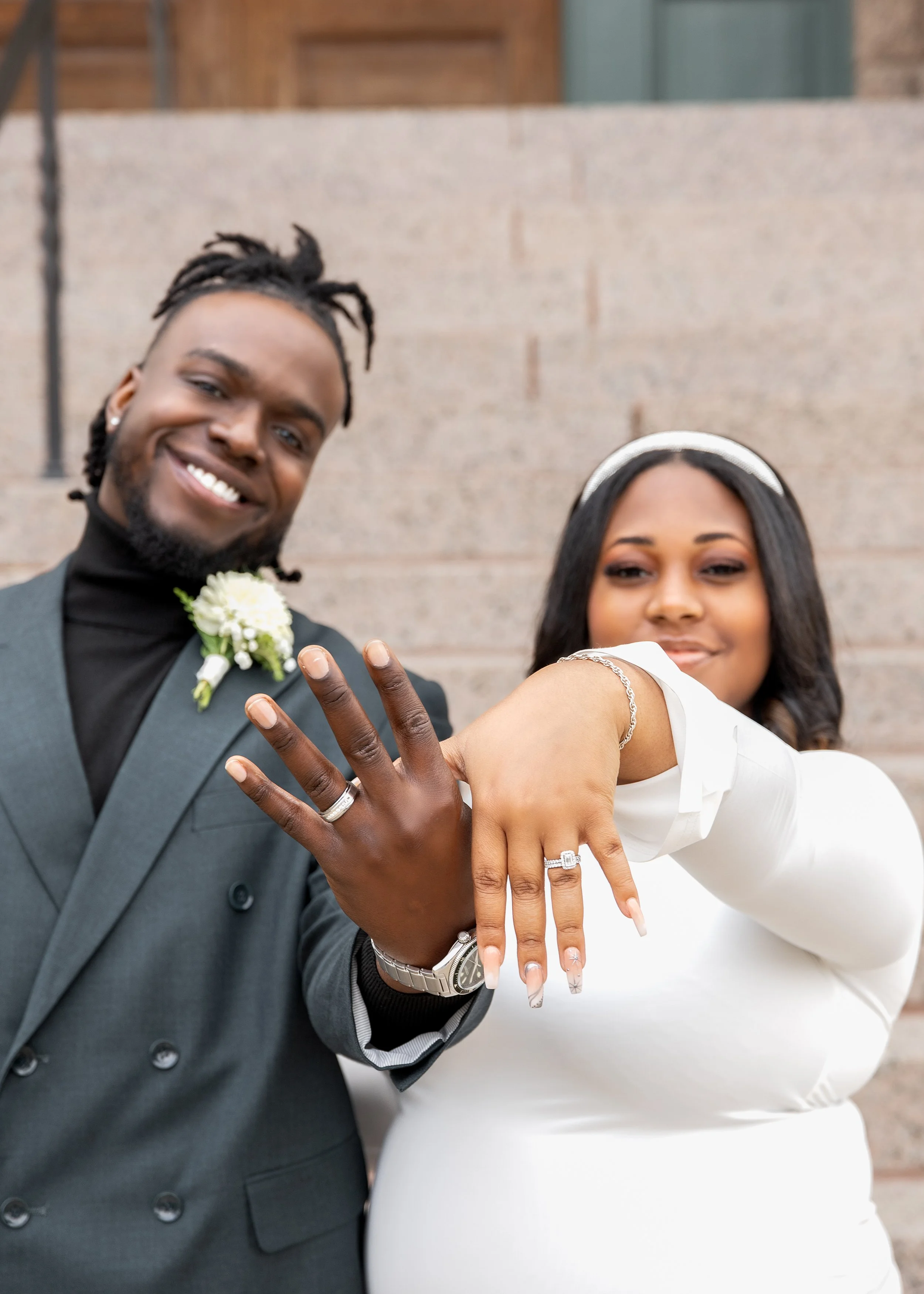 A happy couple showing off rings on their extended hands in front of a brick wall, celebrating their wedding.