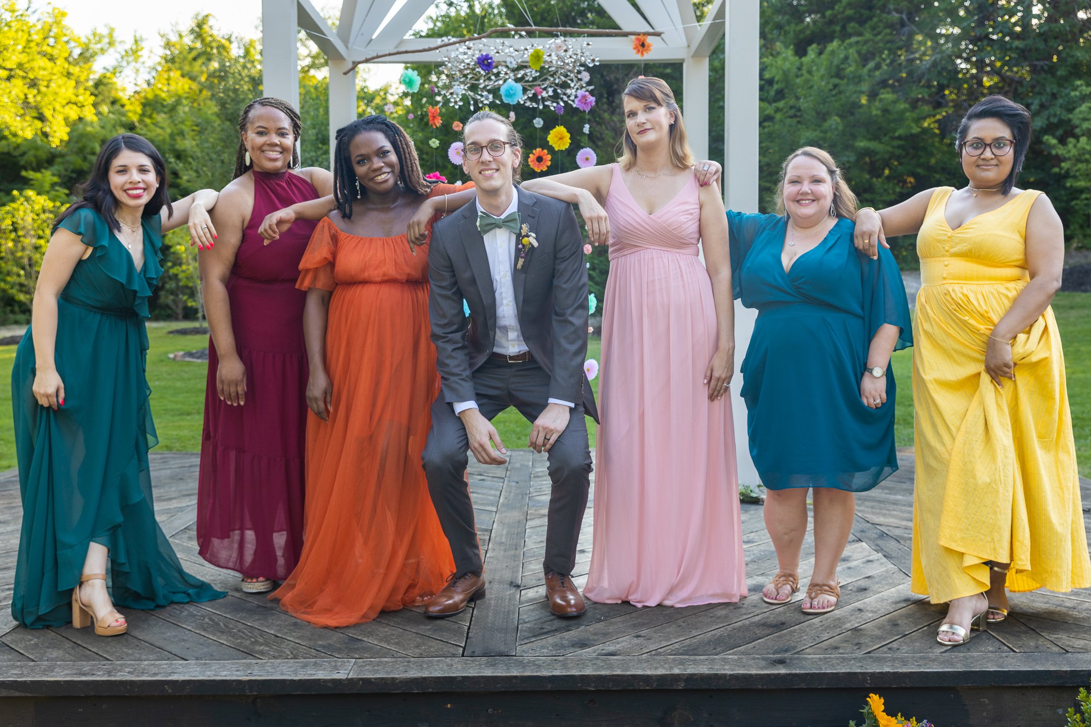Group of seven people dressed in colorful formal dresses and suits standing outdoors on a wooden platform, with arms around each other, smiling at the camera during a celebration or wedding event.