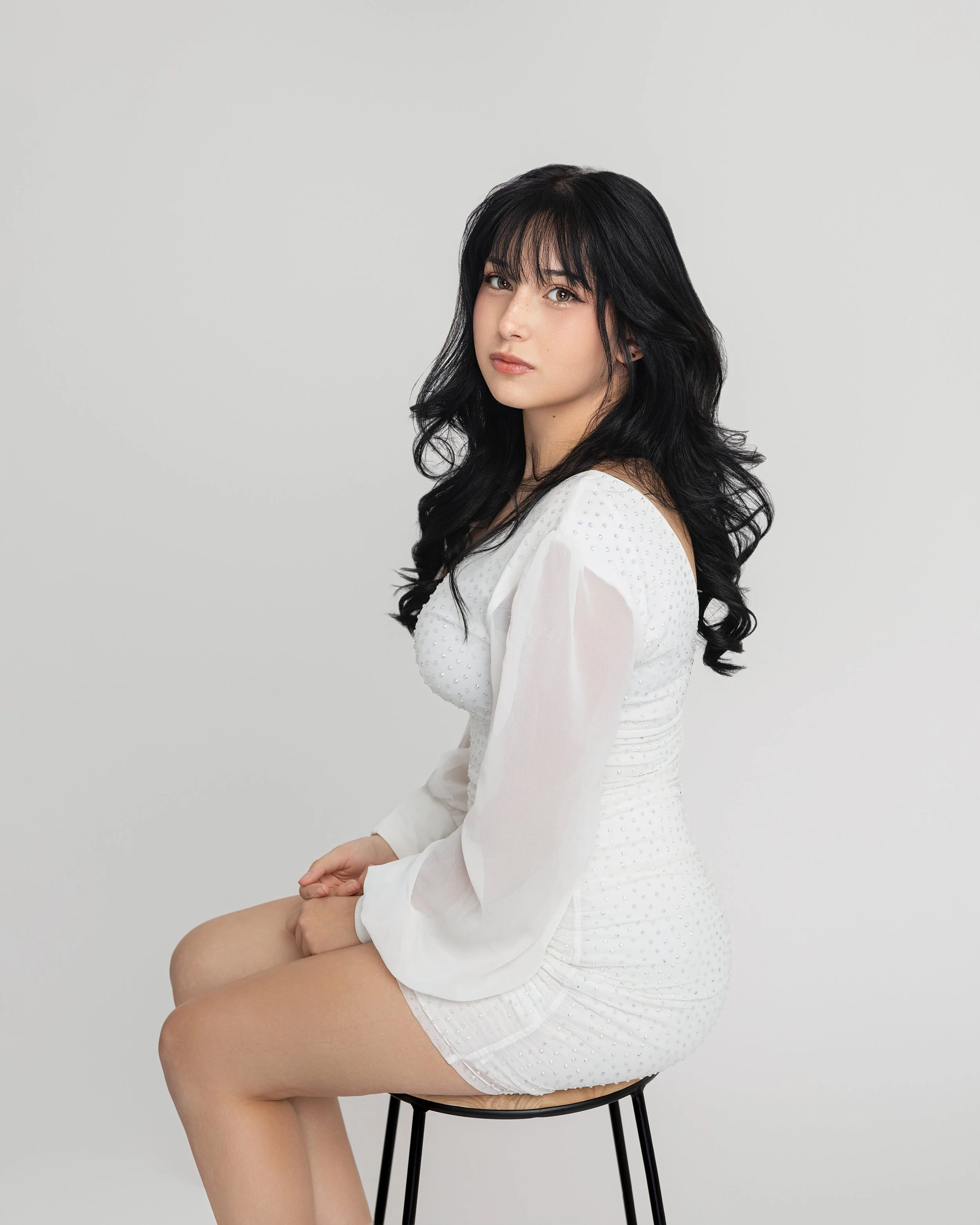 A young woman with long black curly hair, wearing a white dress with sheer long sleeves, sitting on a black stool against a plain light grey background.