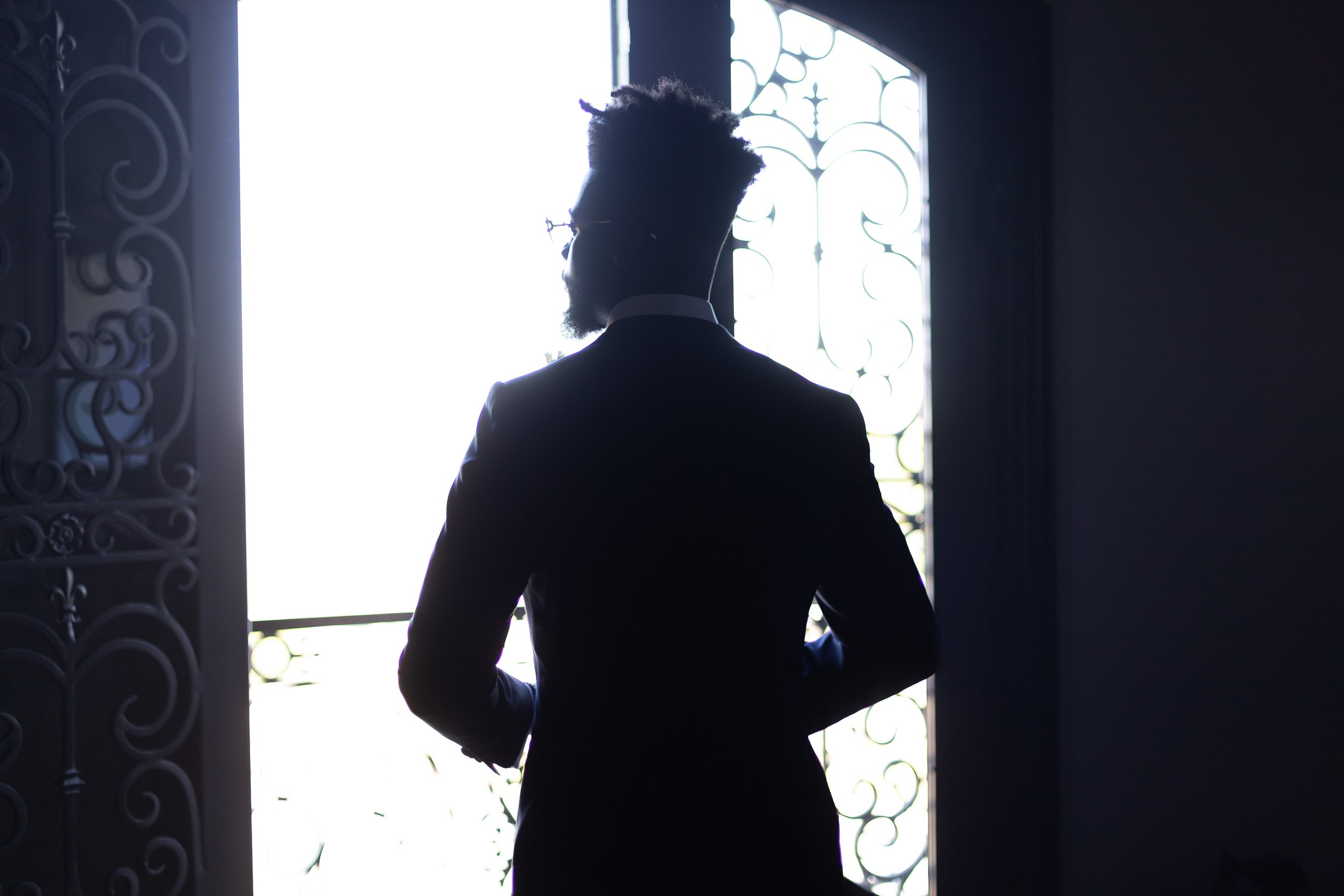 Silhouette of a man in a suit standing in front of a bright window with decorative ironwork.