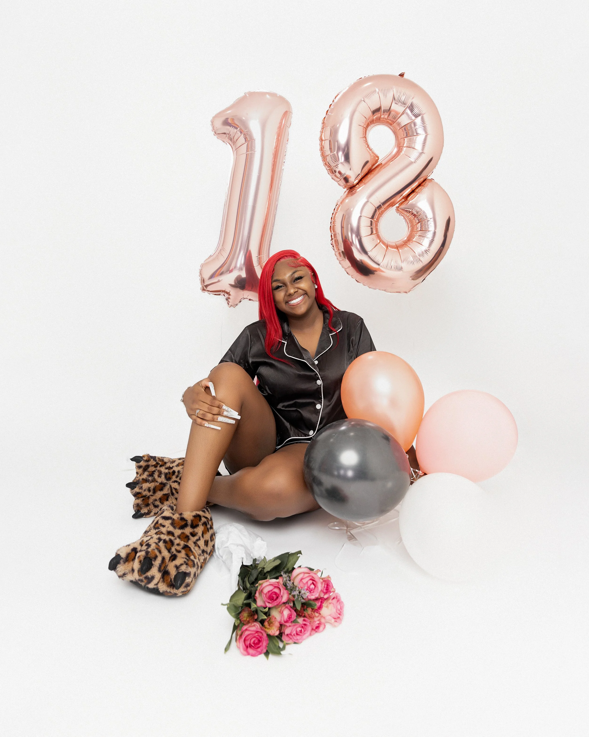 A woman with red hair sitting on the floor, smiling, surrounded by pink, white, and black balloons, a bouquet of pink roses, wearing leopard print slippers and pajamas, celebrating her 18th birthday with large rose gold number balloons in the backgro