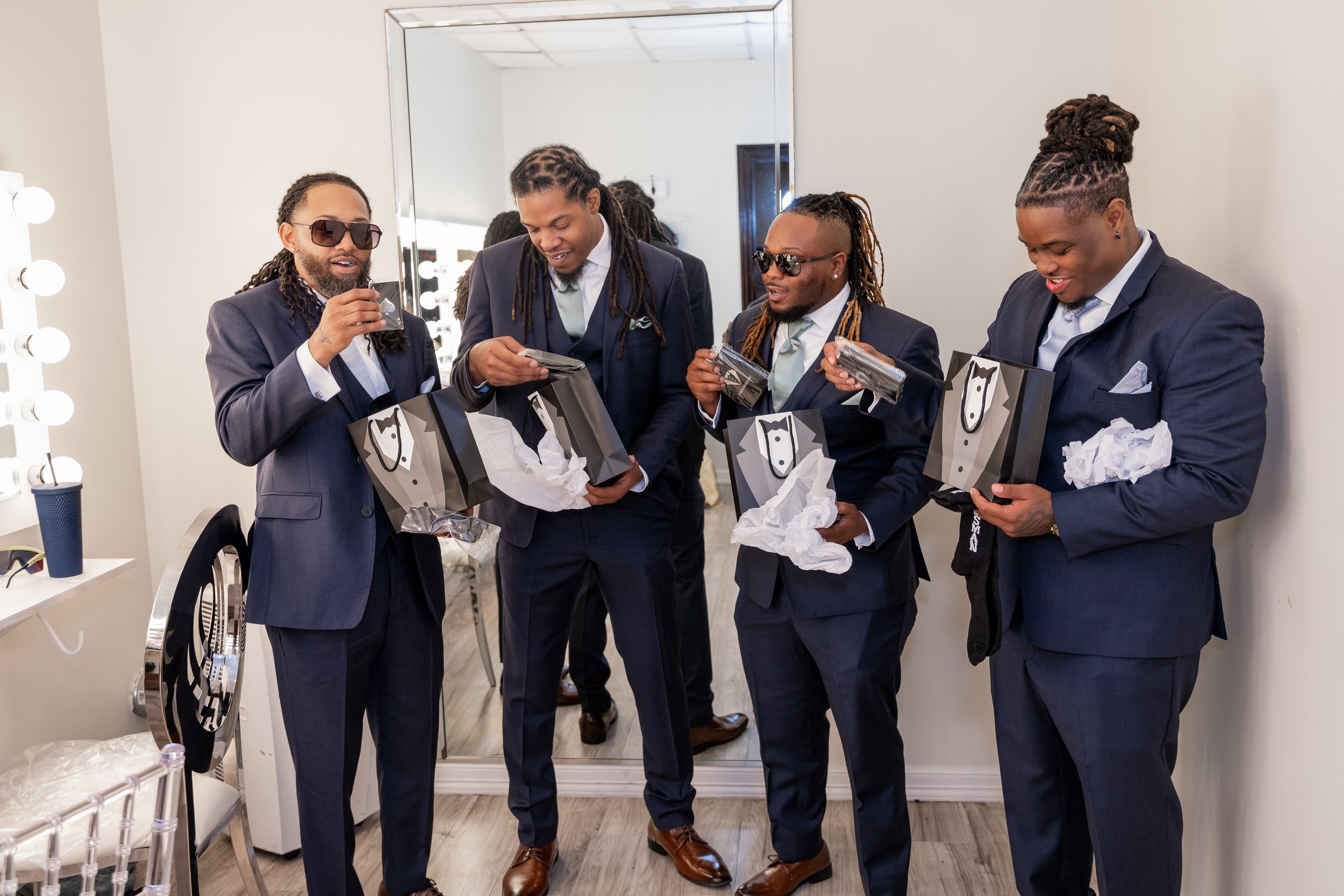 Four men in formal suits with ties and sunglasses, holding gift bags and tissue paper, smiling and enjoying themselves in a dressing room with a mirror and makeup lights.