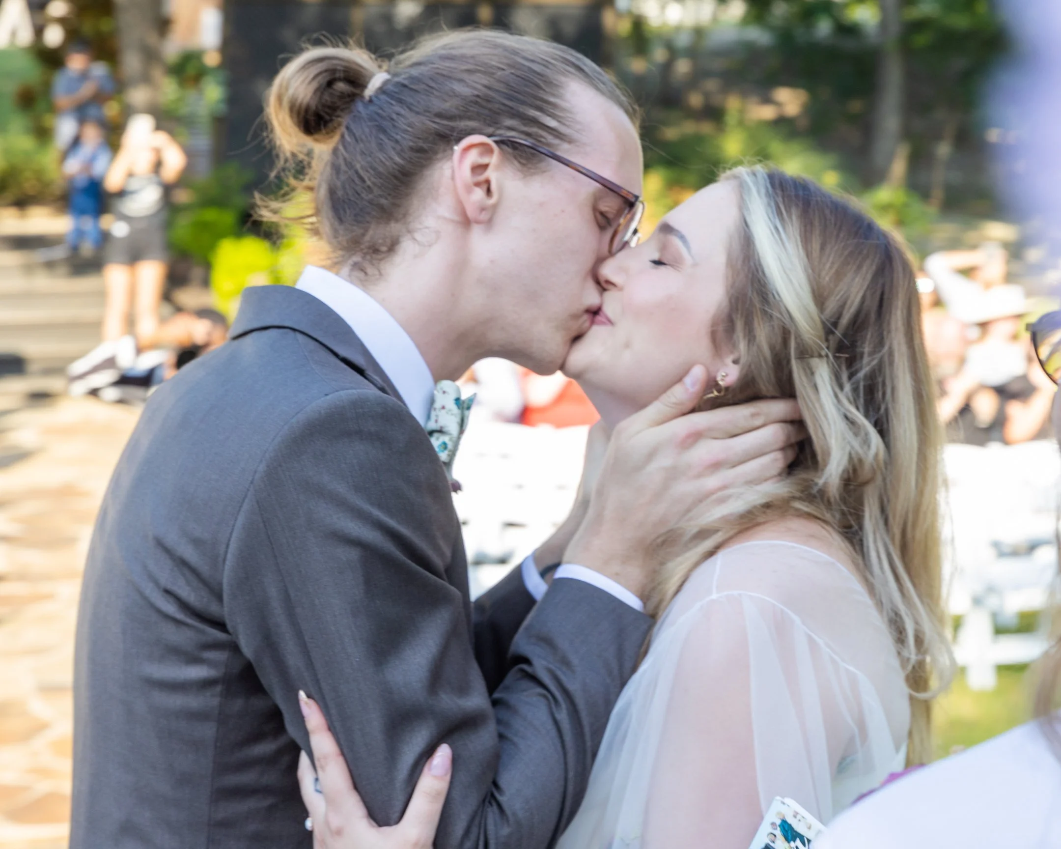 A couple sharing a kiss at an outdoor wedding ceremony, with the groom wearing a suit and glasses, and the bride with long, blonde hair in a semi-sheer dress.