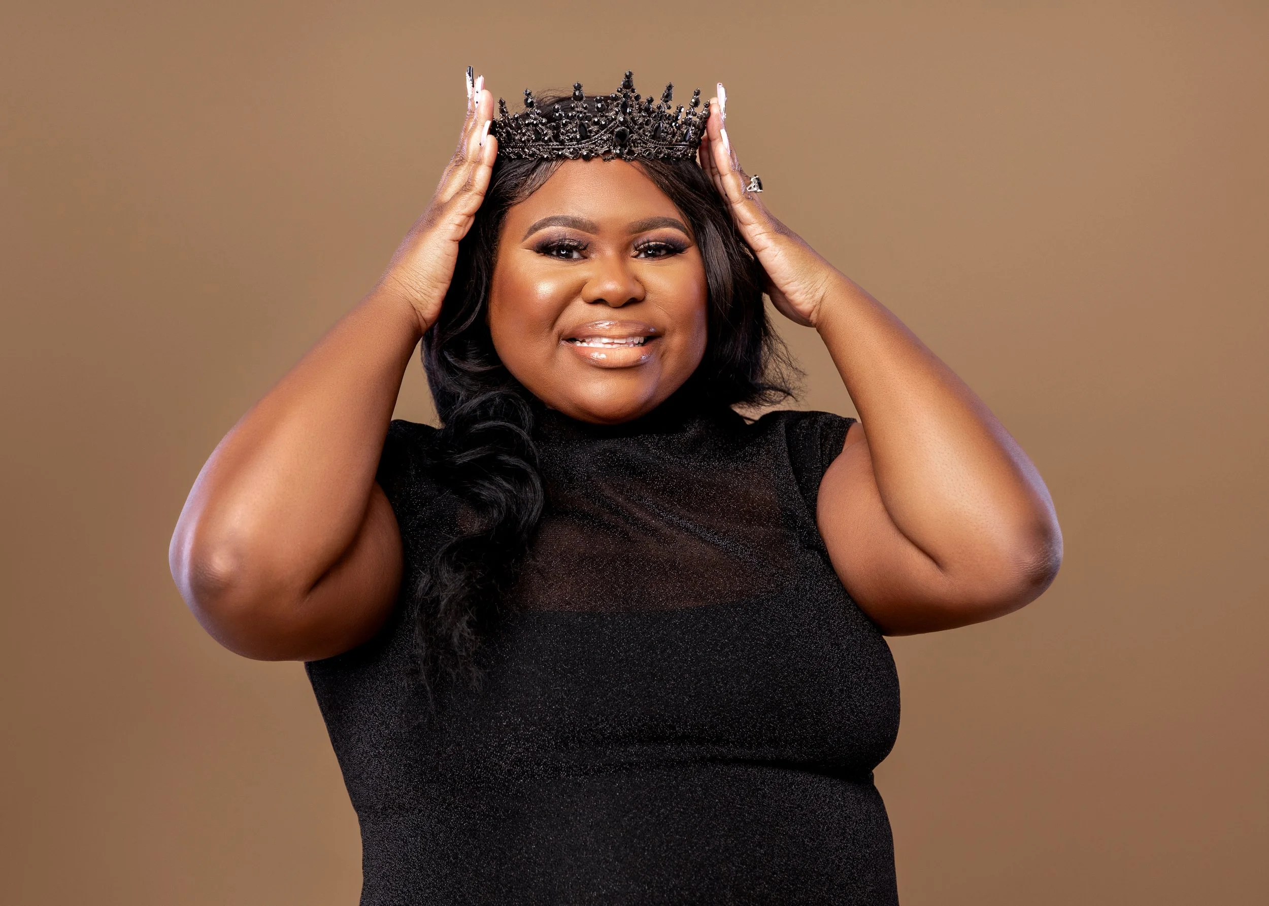 A woman in a black dress wears a crown and holds it on her head, smiling confidently against a plain background.