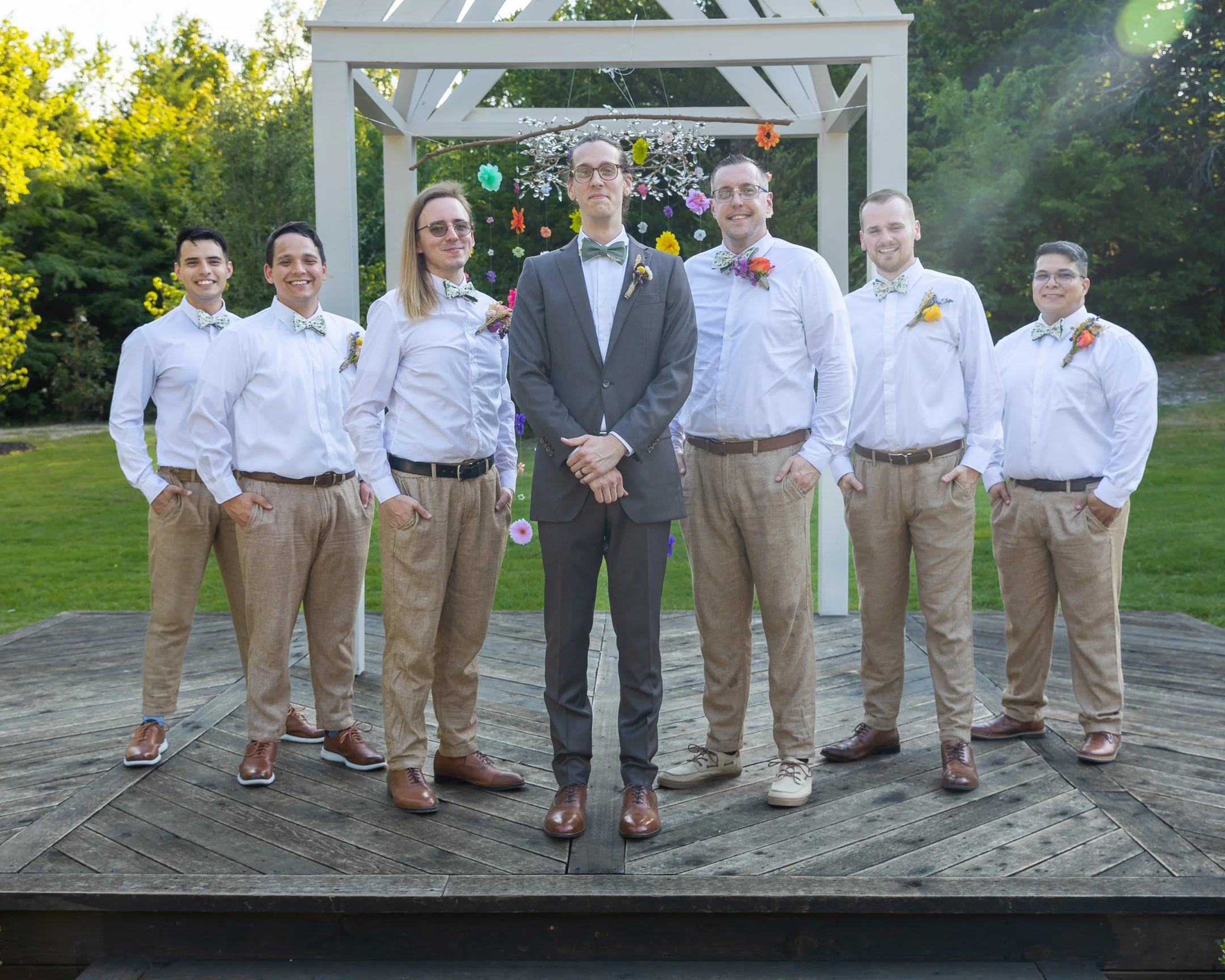 Groom and six groomsmen standing outdoors on a wooden platform, with a white arch decorated with colorful flowers behind them, surrounded by green trees.