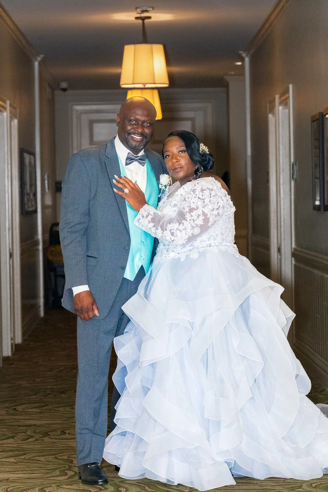 A bride and groom dressed in wedding attire posing together in an indoor setting, with the bride wearing a white wedding gown with ruffled layers and lace top, and the groom in a gray suit with a light turquoise vest and bowtie. They are standing clo