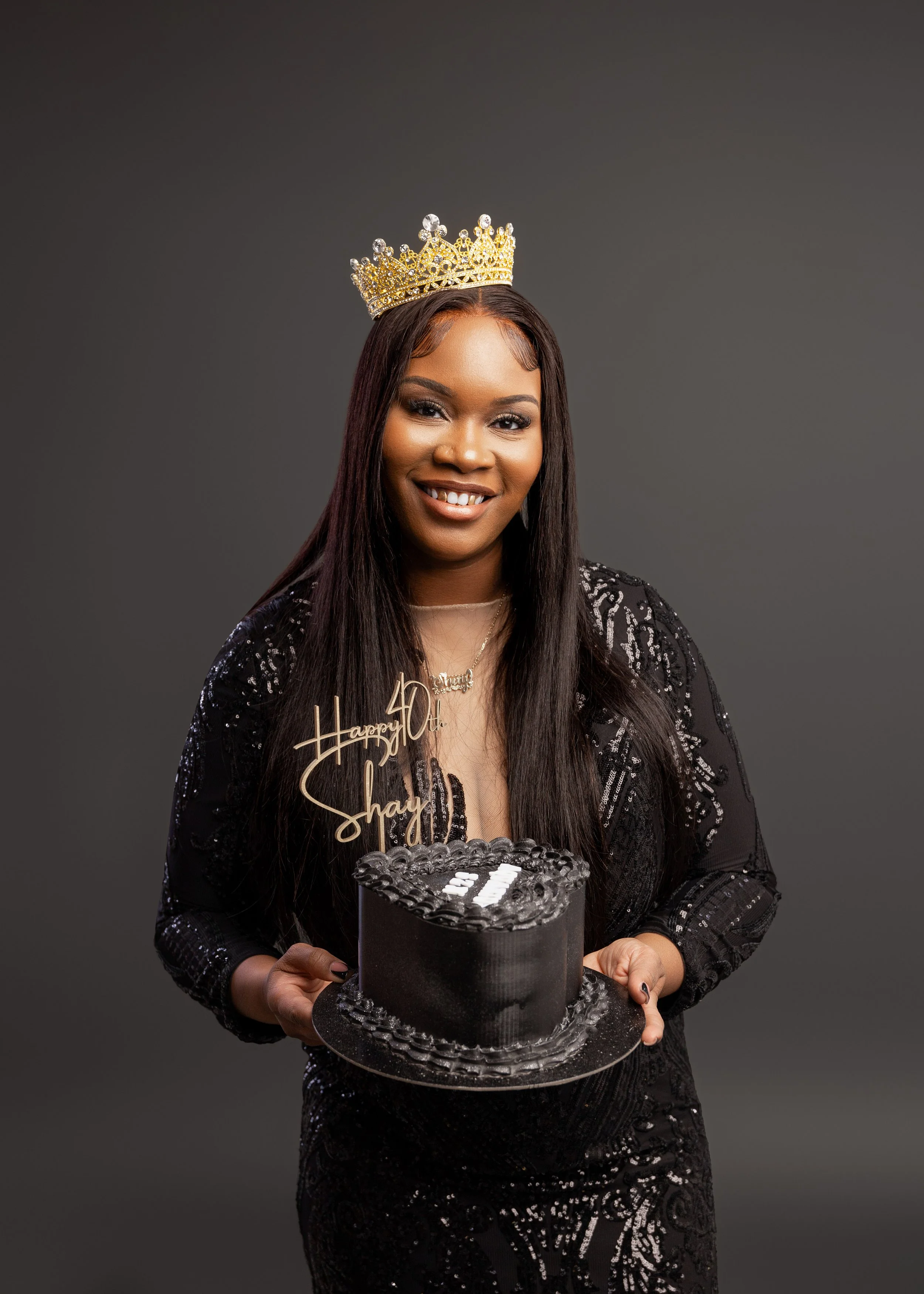 A woman wearing a tiara and black sequined dress is holding a black birthday cake with a "40" topper and a "Happy 40th Shay" sign, smiling against a dark gray background.