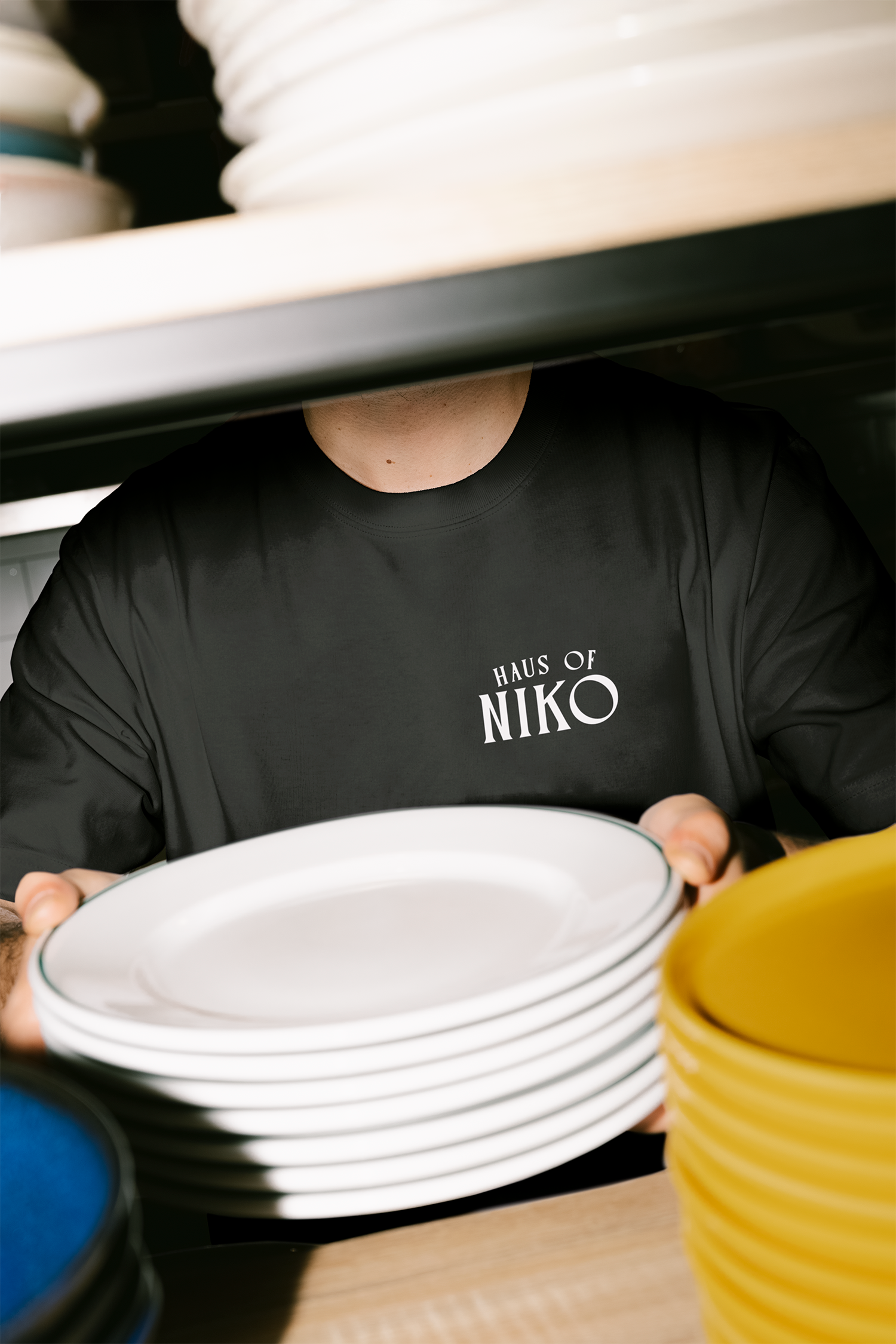Person wearing a black "Haus of Niko" t-shirt is reaching for a white bowl on a table among stacked yellow and blue cans.