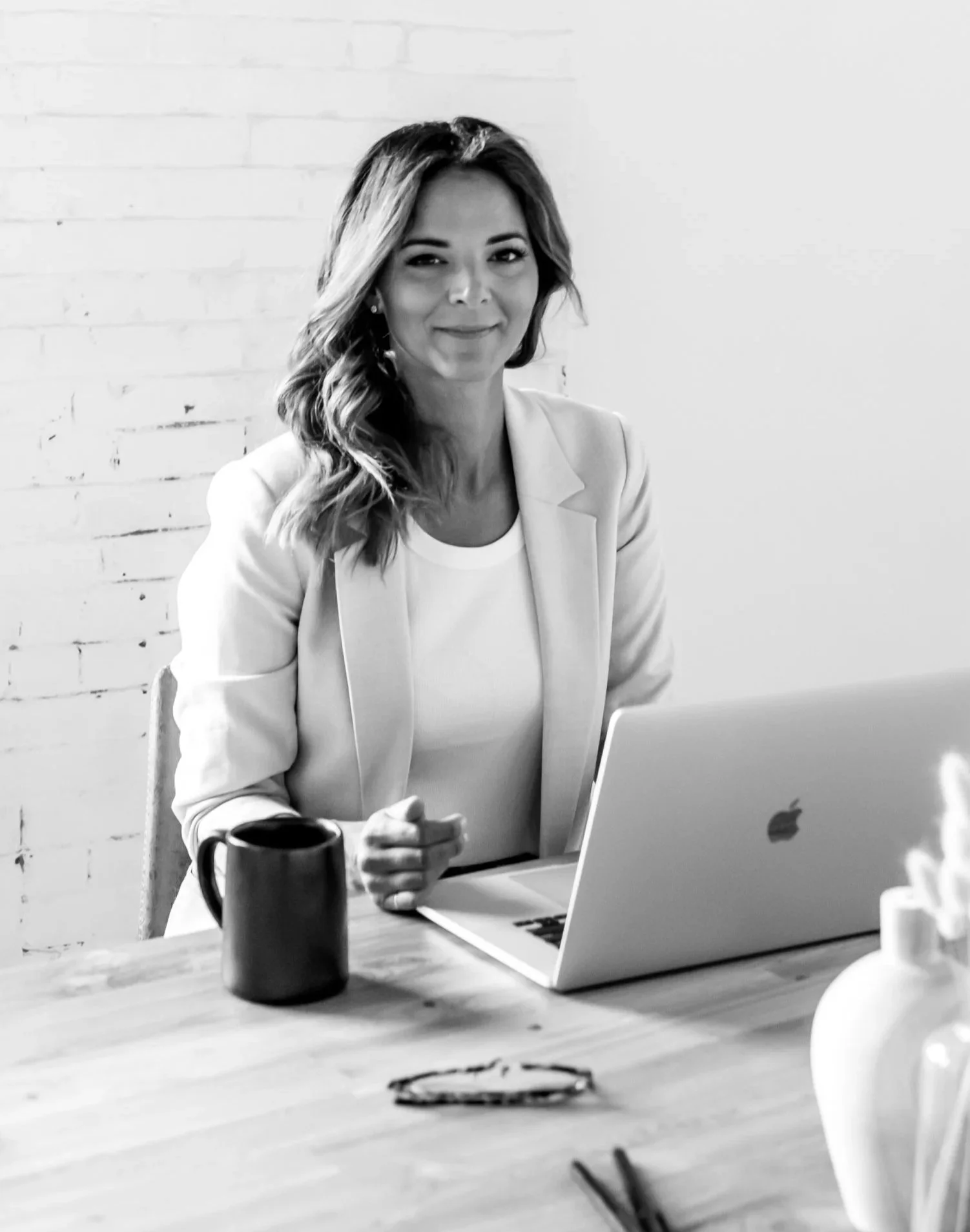 Black and white photo of a woman sitting at a table with a laptop, coffee mug, and other items, smiling at the camera.