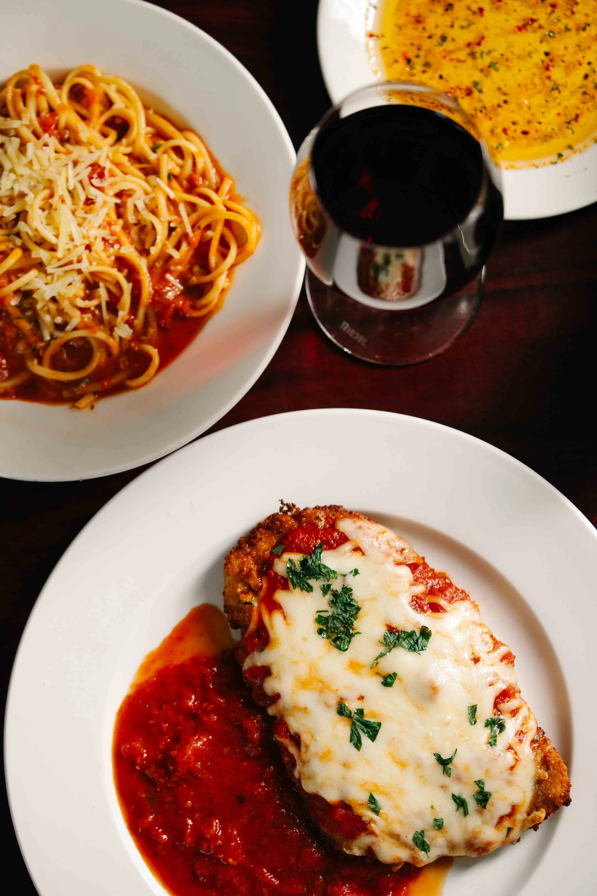 A plate of baked lasagna topped with melted cheese and chopped herbs, served with tomato sauce. In the background, a bowl of spaghetti with tomato sauce and grated cheese, a bowl of soup, and a glass of red wine.