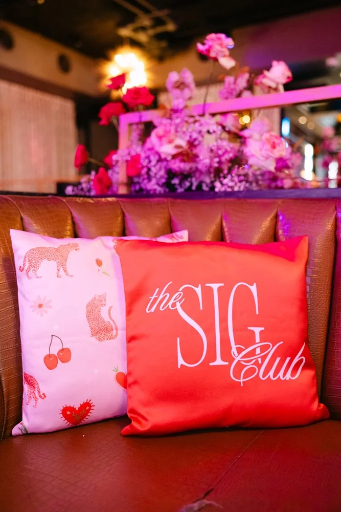 Decorative throw pillows on a brown leather sofa, one with a pink leopard print and cherries, and the other with the words 'the SIG Club' in white on an orange background, in a dimly lit lounge with pink and purple floral arrangements in the backgrou