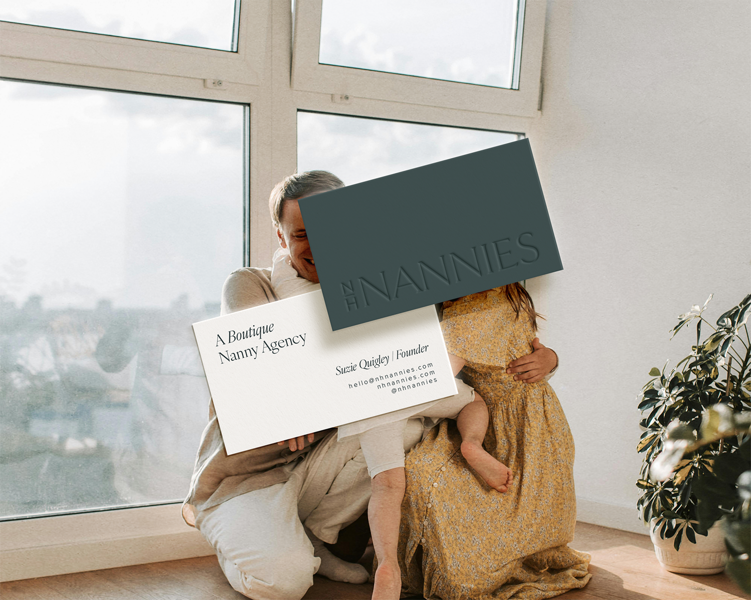 A man and a young girl hugging indoors near a large window with cloudy sky view, holding business cards for The Nannies and Nanniez, a boutique nanny agency.