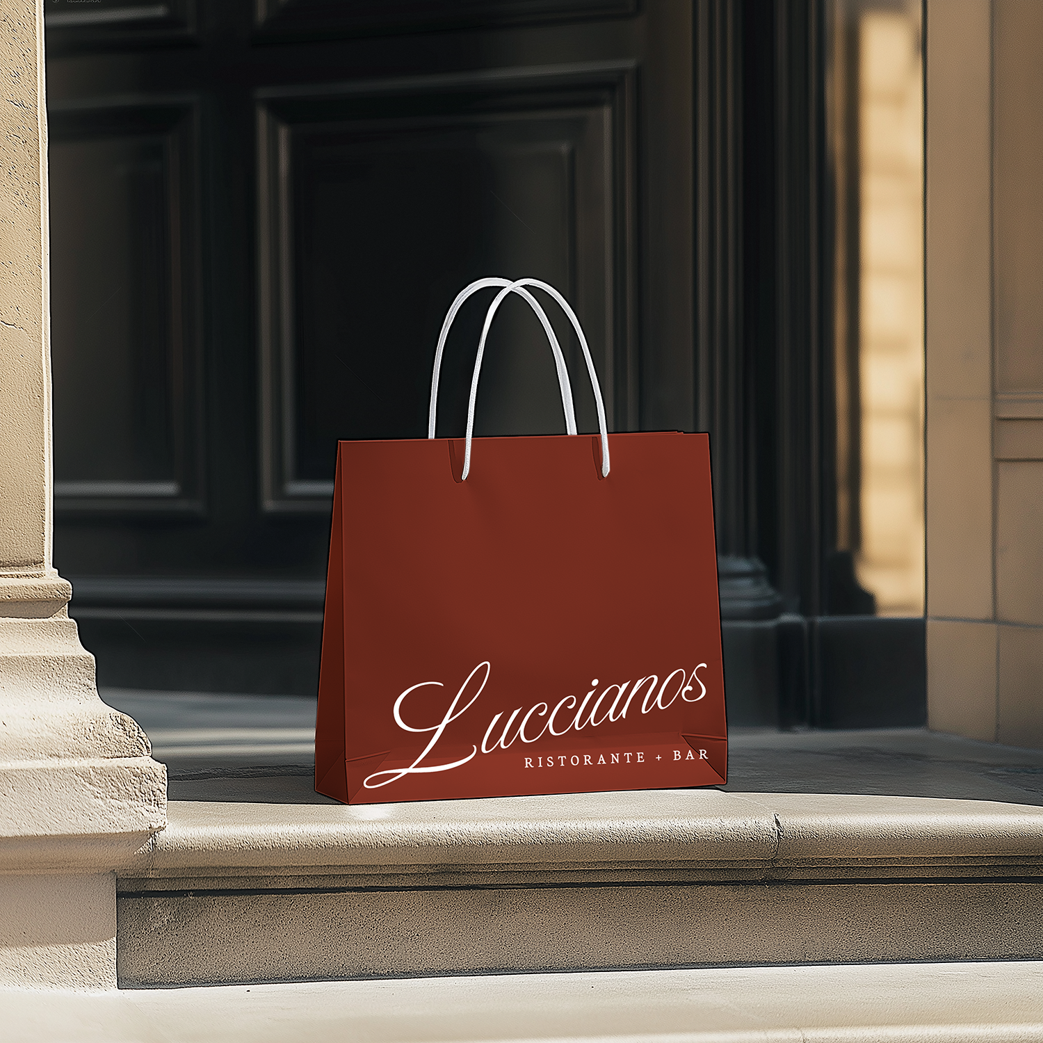 A red paper bag with white handles sitting on a stone ledge in front of a black door, with the label "Luccianos Ristorante + Bar" written on it.
