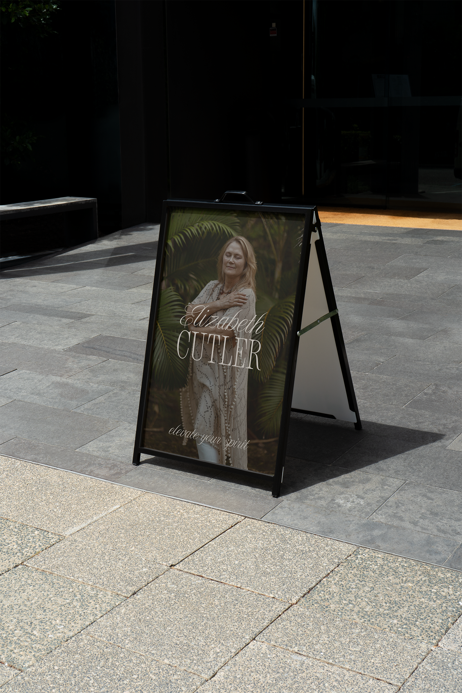 A standing signboard outdoors featuring a woman with blonde hair with her eyes closed, wearing a beige patterned coat, surrounded by large green leaves, with text that reads "Elizabeth Cutler" and "elevate your spirit" on the sign.