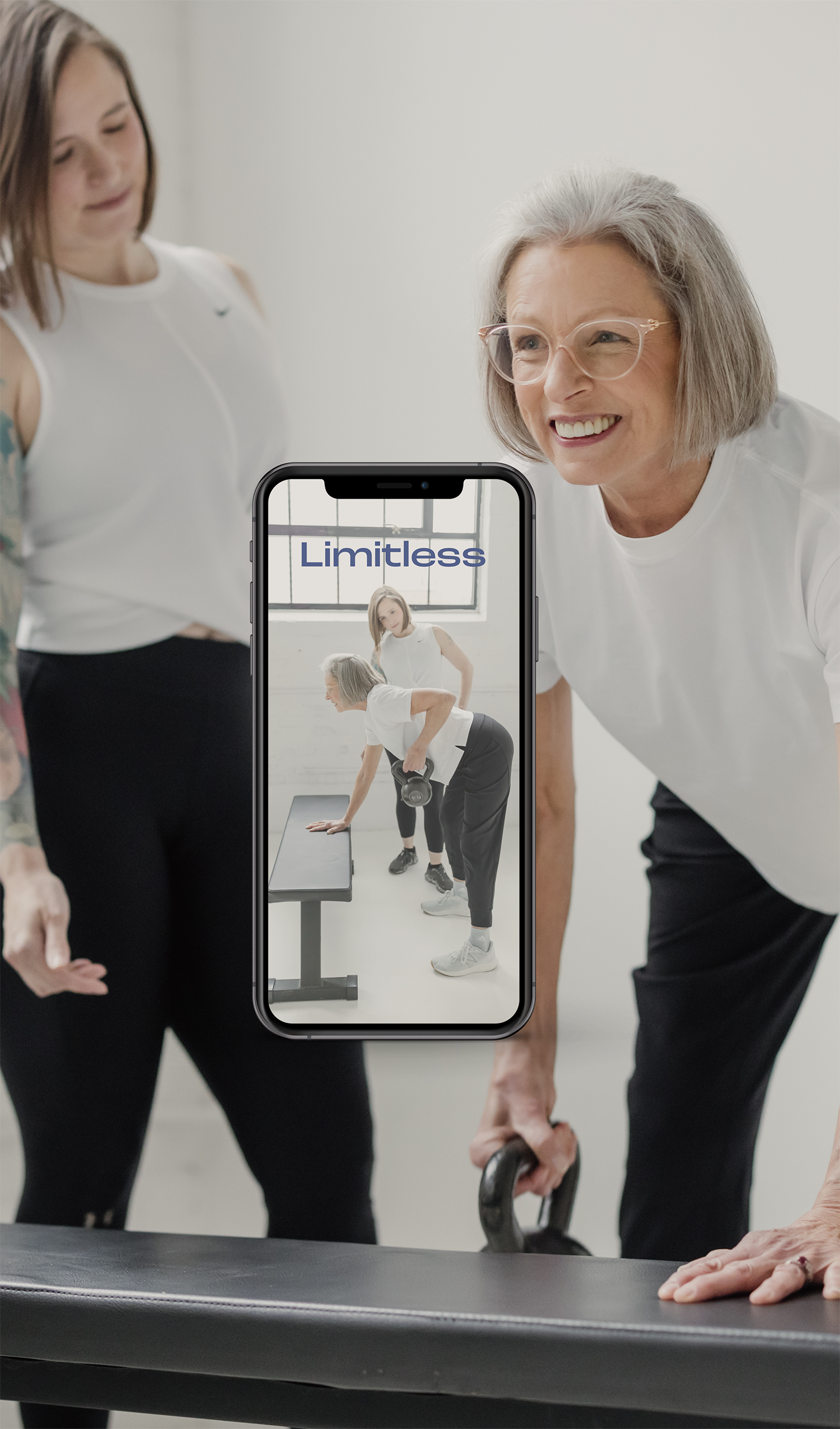 Older woman and young woman exercising in a fitness class, using a kettlebell and a workout bench, with a smartphone capturing the scene with the word 'Limitless' displayed.