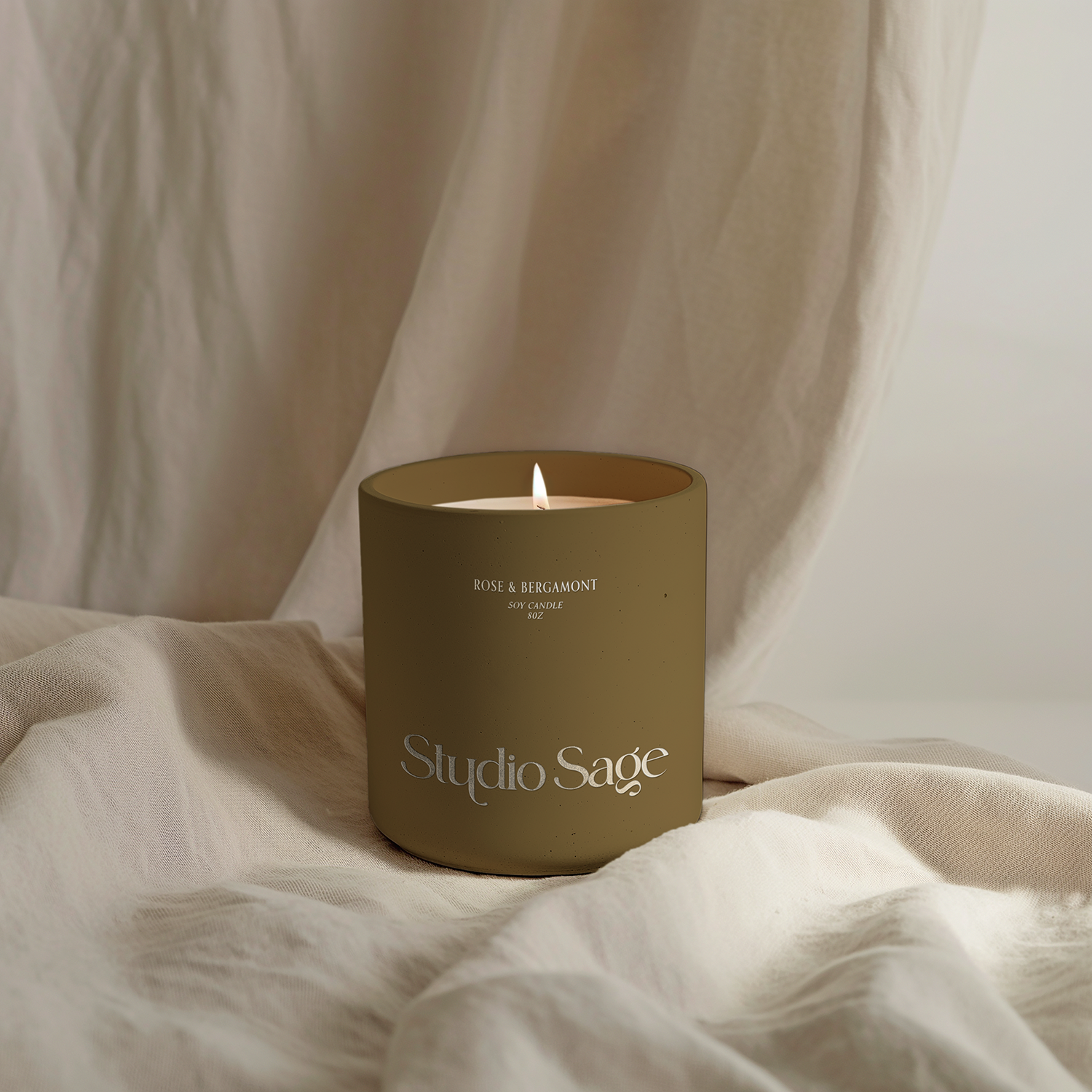 A beige soy candle labeled 'Studio Sage' with a lit wick, placed on cream-colored fabric with a soft drape background.