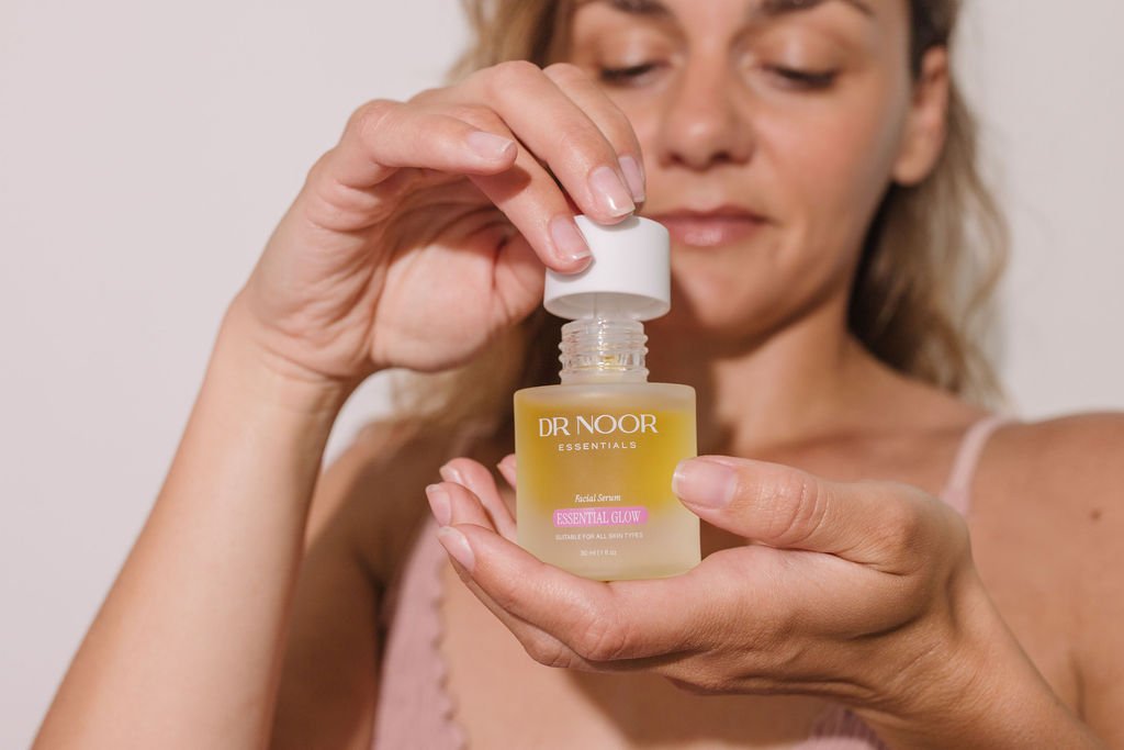 A woman holding a small clear bottle of facial serum labeled 'DR NOOR ESSENTIALS Face Serum Essential Glow' and opening it.