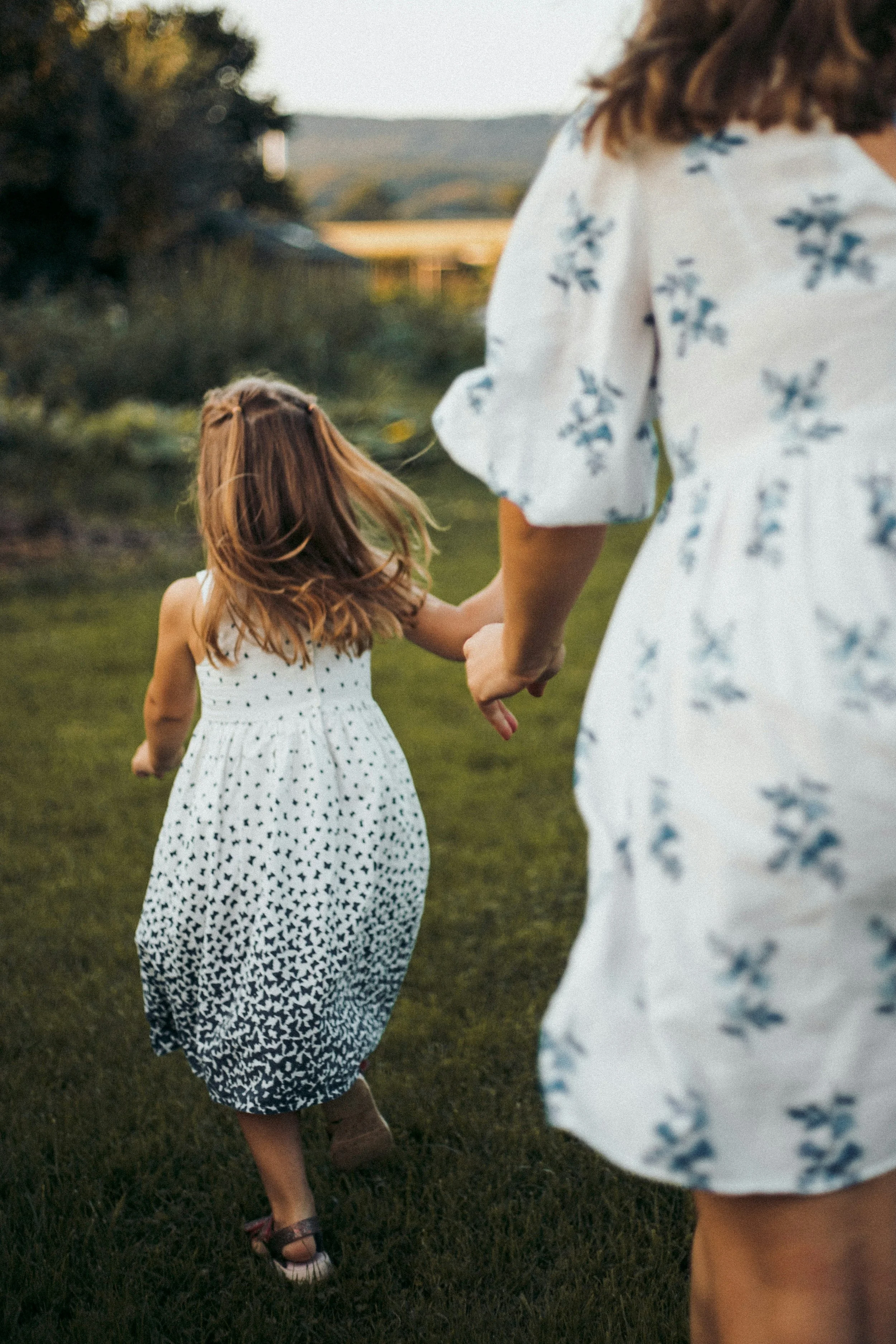 A woman and a child holding hands and walking on a grassy field during sunset, with trees and distant hills in the background.