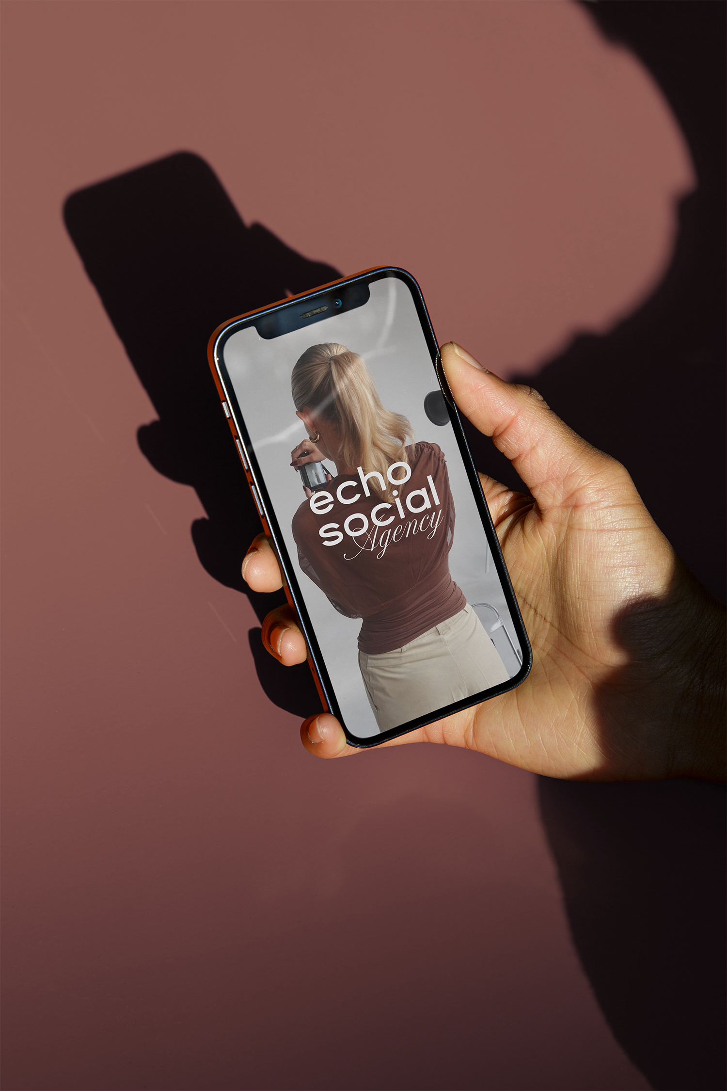 A person's hand holding a smartphone displaying a photo of a woman with blonde hair, wearing a brown top and beige skirt, with the words 'echo social Agency' on the screen. The background is pink with a shadow of the phone and hand.