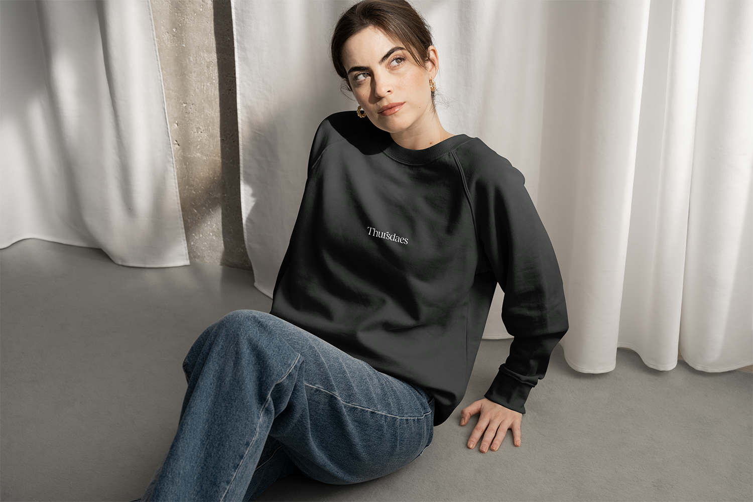 A woman with dark hair, fair skin, and gold hoop earrings sitting on the floor near white curtains, wearing a black sweatshirt with the word 'Thürdaes' embroidered on it and blue jeans.