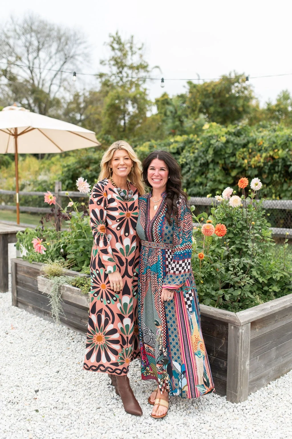 Two women standing outdoors in a garden with blooming flowers, smiling at the camera. One woman is wearing a colorful floral dress with long sleeves, and the other is in a vibrant patchwork dress. There is a white umbrella and string lights in the background.