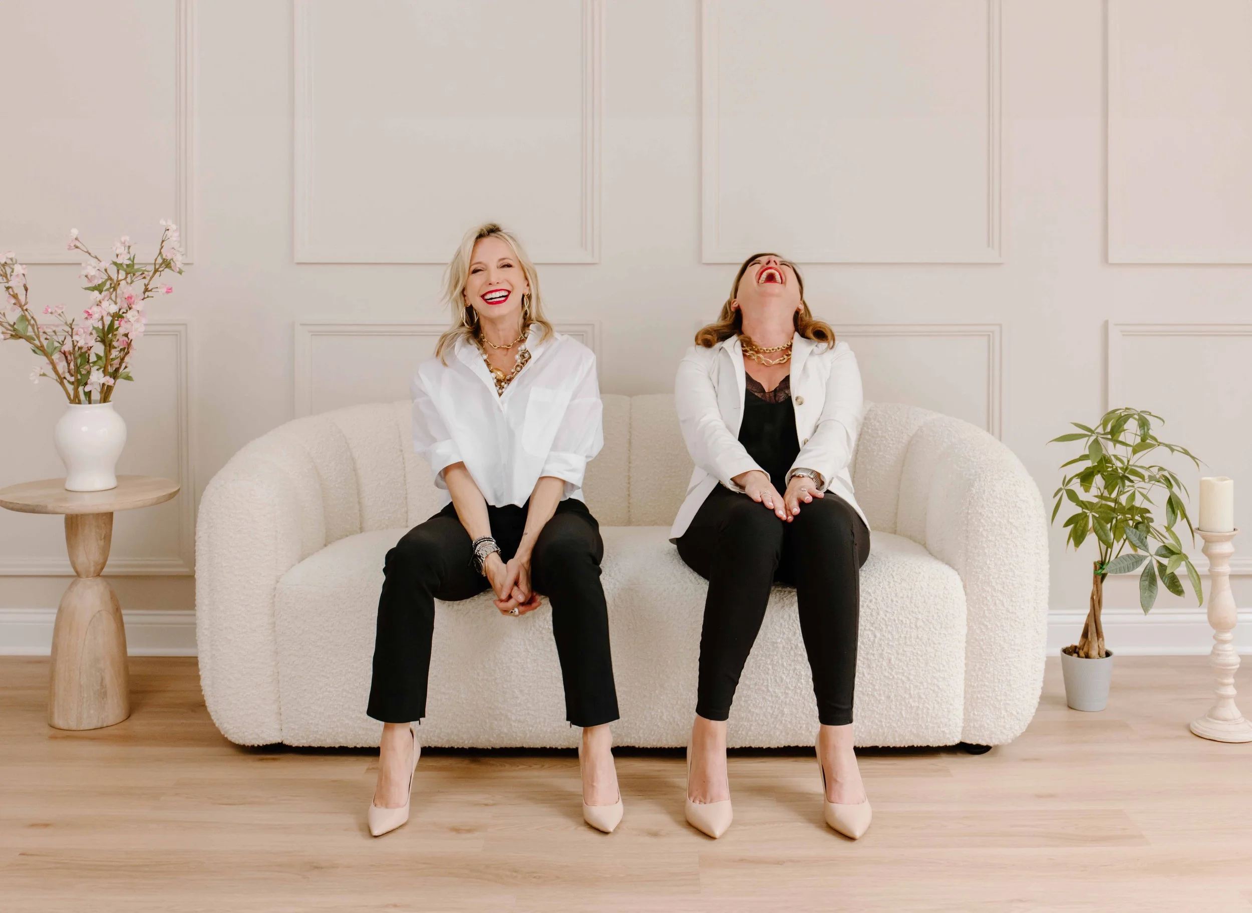 Two women sitting on a white sofa in a minimalist room, laughing and enjoying each other's company. They are dressed in white tops and black pants, wearing high heels, with pink lipstick and jewelry. There are plants and flowers around them, including a vase with pink blossoms on a wooden side table.