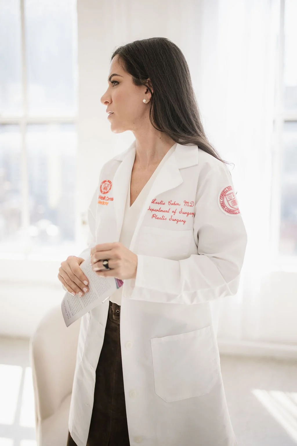 A female doctor wearing a white lab coat with embroidered text, standing indoors near windows, holding a paper and looking to her left.