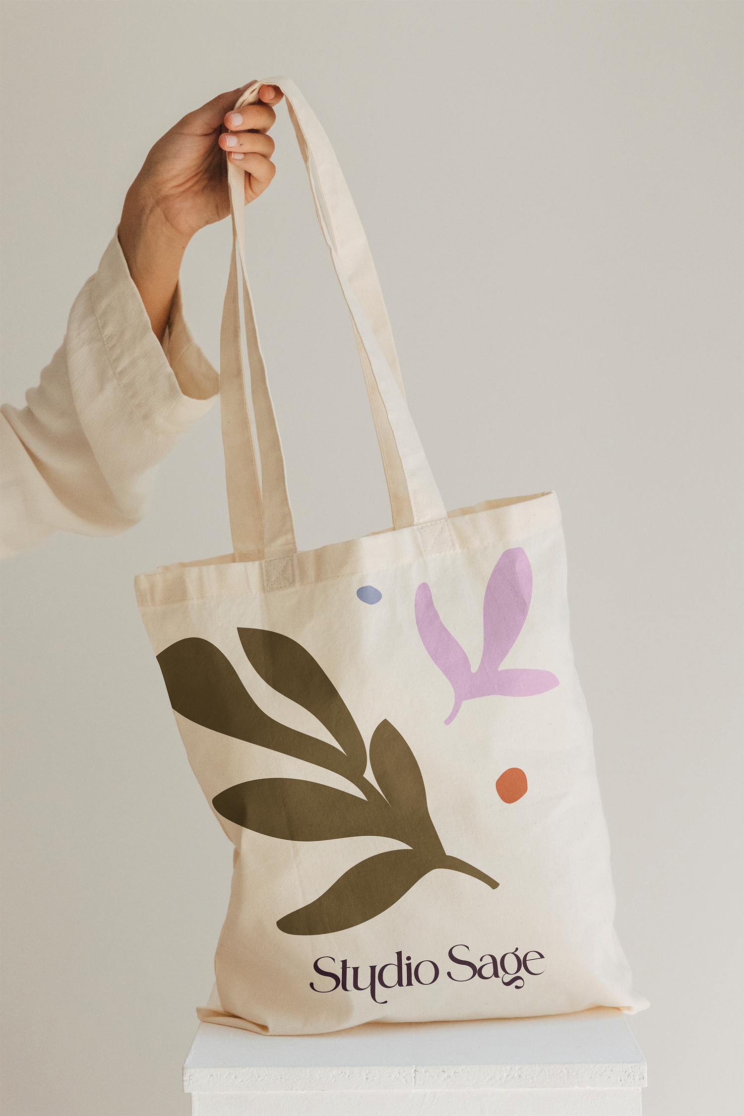 A cream-colored tote bag with colorful abstract shapes and the text 'Studio Sage' printed on it, held by a person's hand against a plain background.