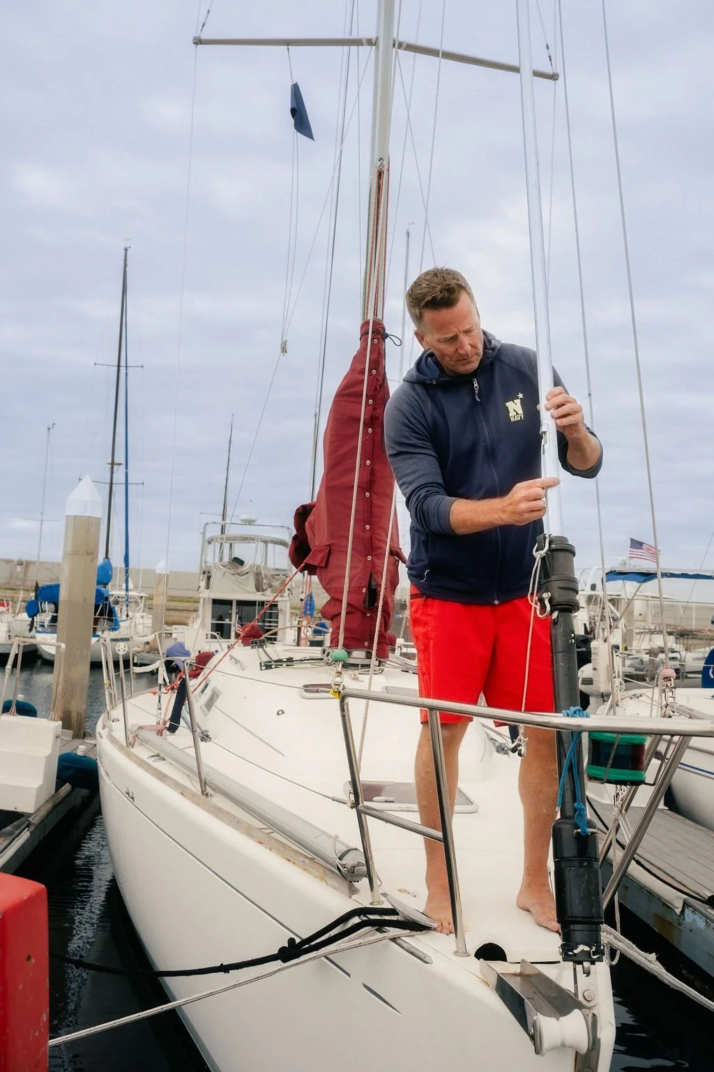 A man standing on a sailboat adjusting a sail at a marina with other boats docked in the background.
