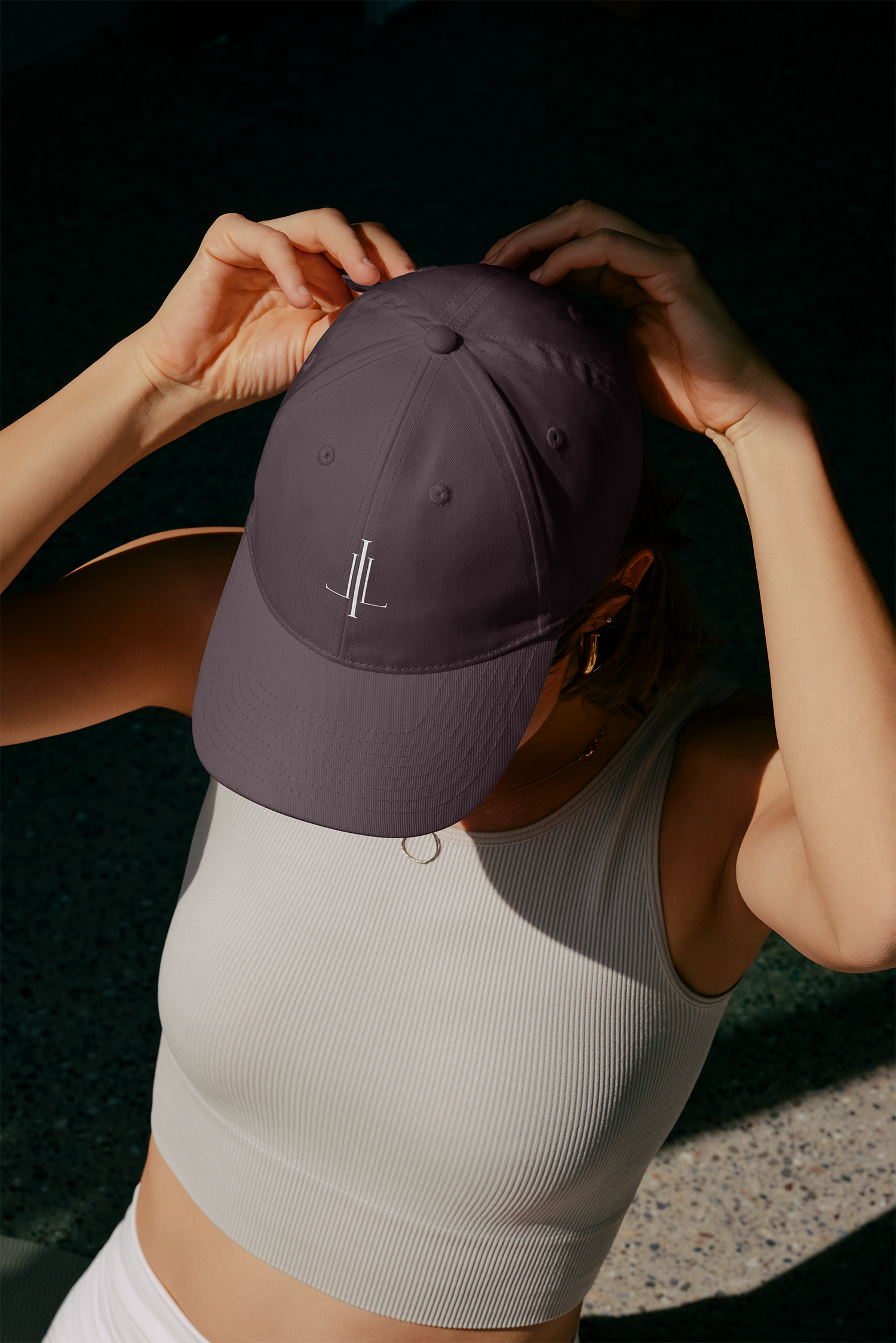 A person wearing a beige athletic tank top and white shorts, holding a purple cap with the initials 'JL' on the front, with a dark background and shadows.