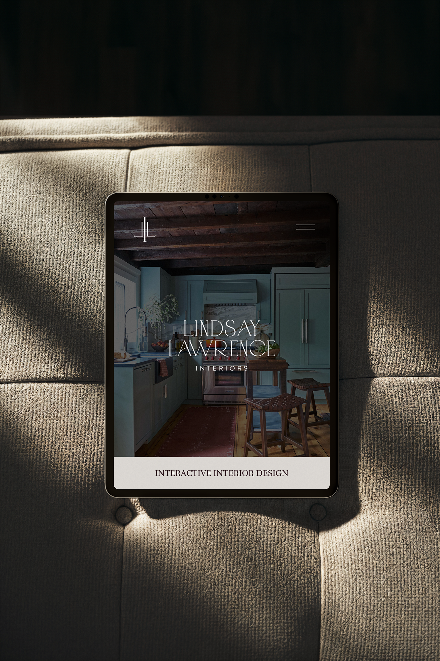 Digital tablet displaying an interior design website with a kitchen image and the text 'Lindsay Lawrence Interiors' and 'Interactive Interior Design' over a brown, broiled kitchen with cabinets and chairs.