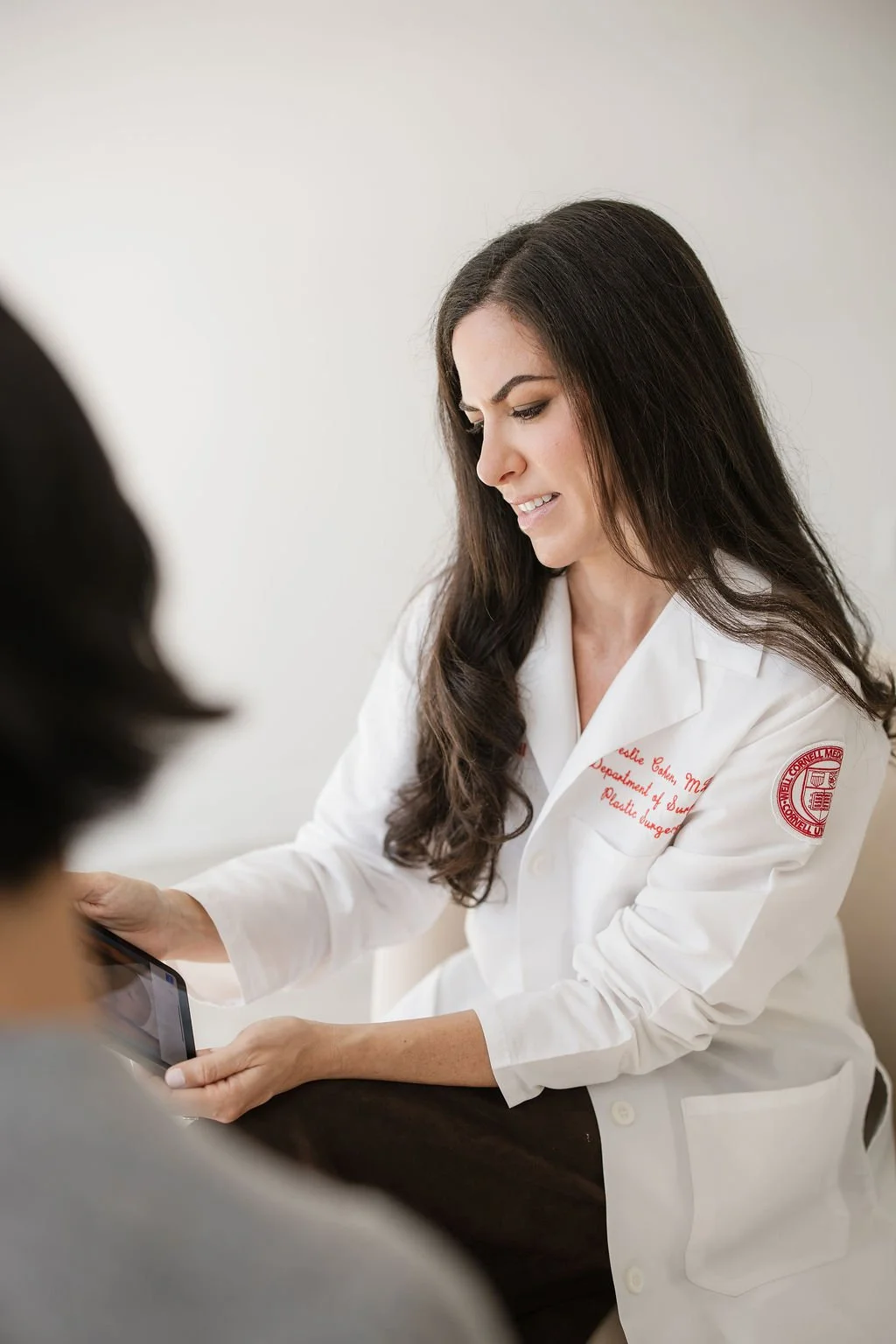 A female doctor with long dark hair, wearing a white lab coat, is showing something on a tablet to a patient in a clinical setting.