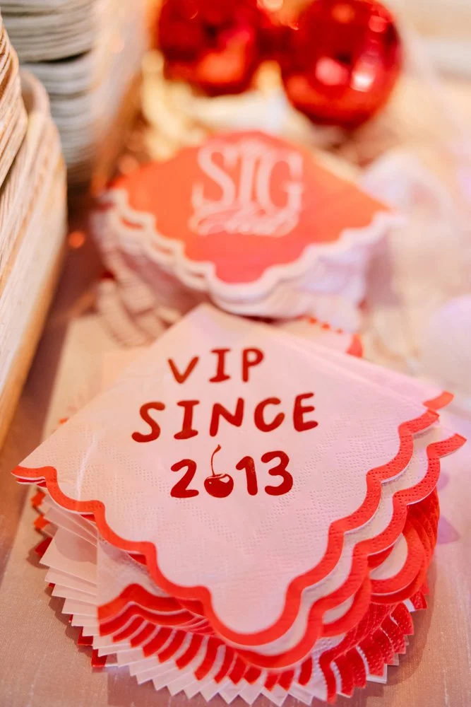 Stack of paper napkins with 'VIP SINCE 2013' written in red and a cherry illustration, on a table with festive decorations in the background.
