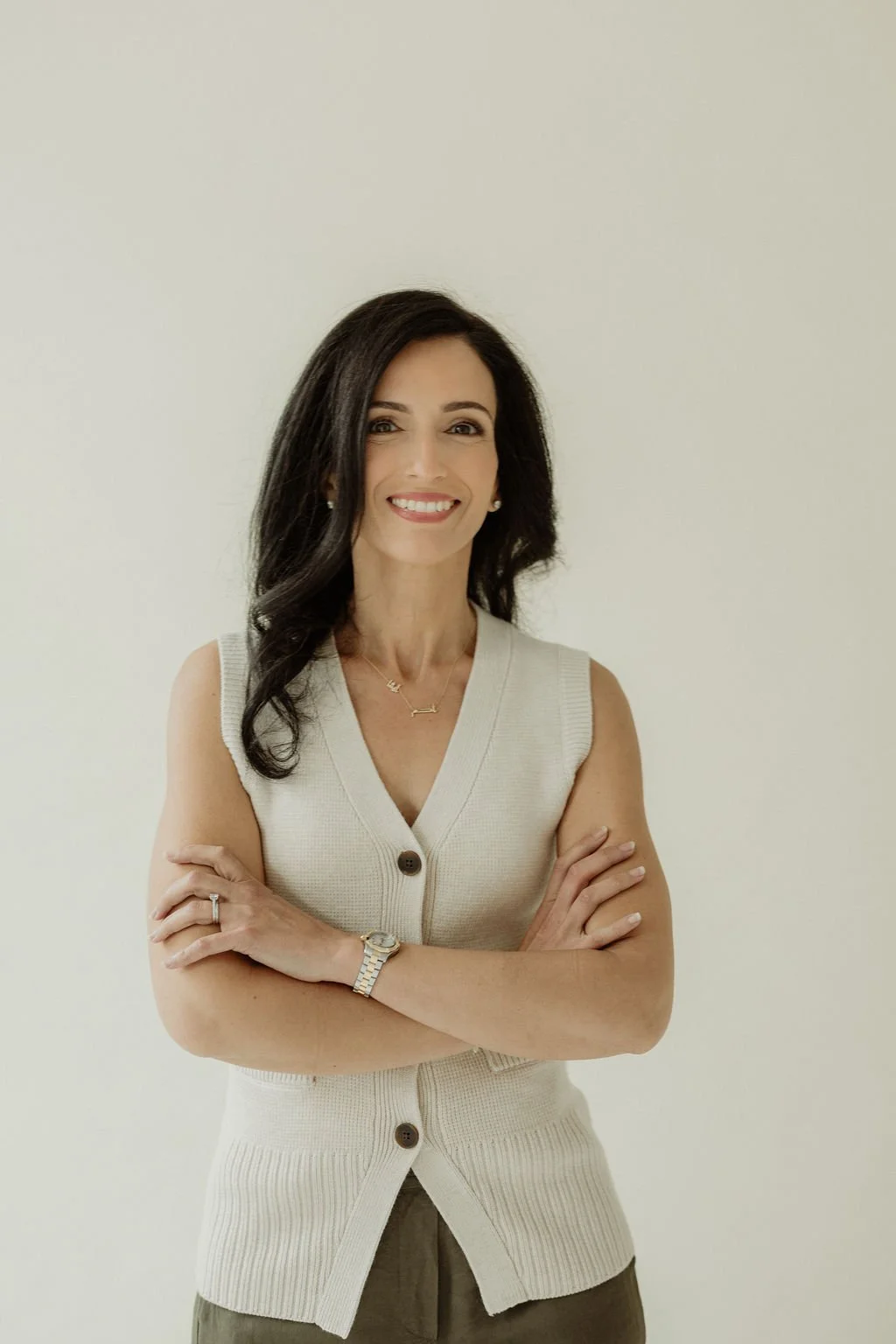 A woman with dark, wavy hair smiling, wearing a sleeveless beige cardigan, jewelry, and standing against a plain light background.