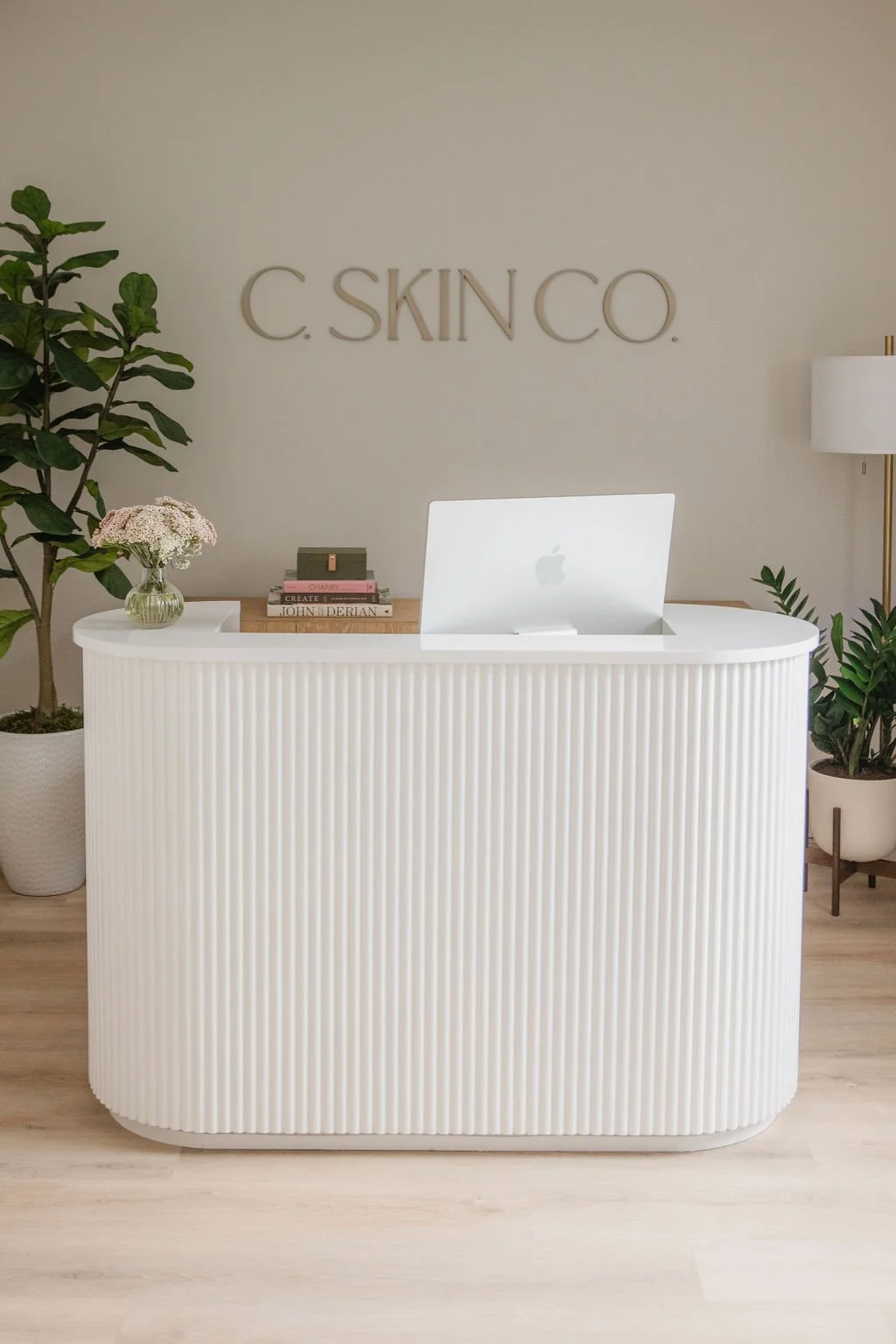 Modern reception desk with white ribbed front, a MacBook, a small stack of books, a glass vase with pink flowers, and potted plants, with a sign on the wall reading 'C. SKINCO' behind it.