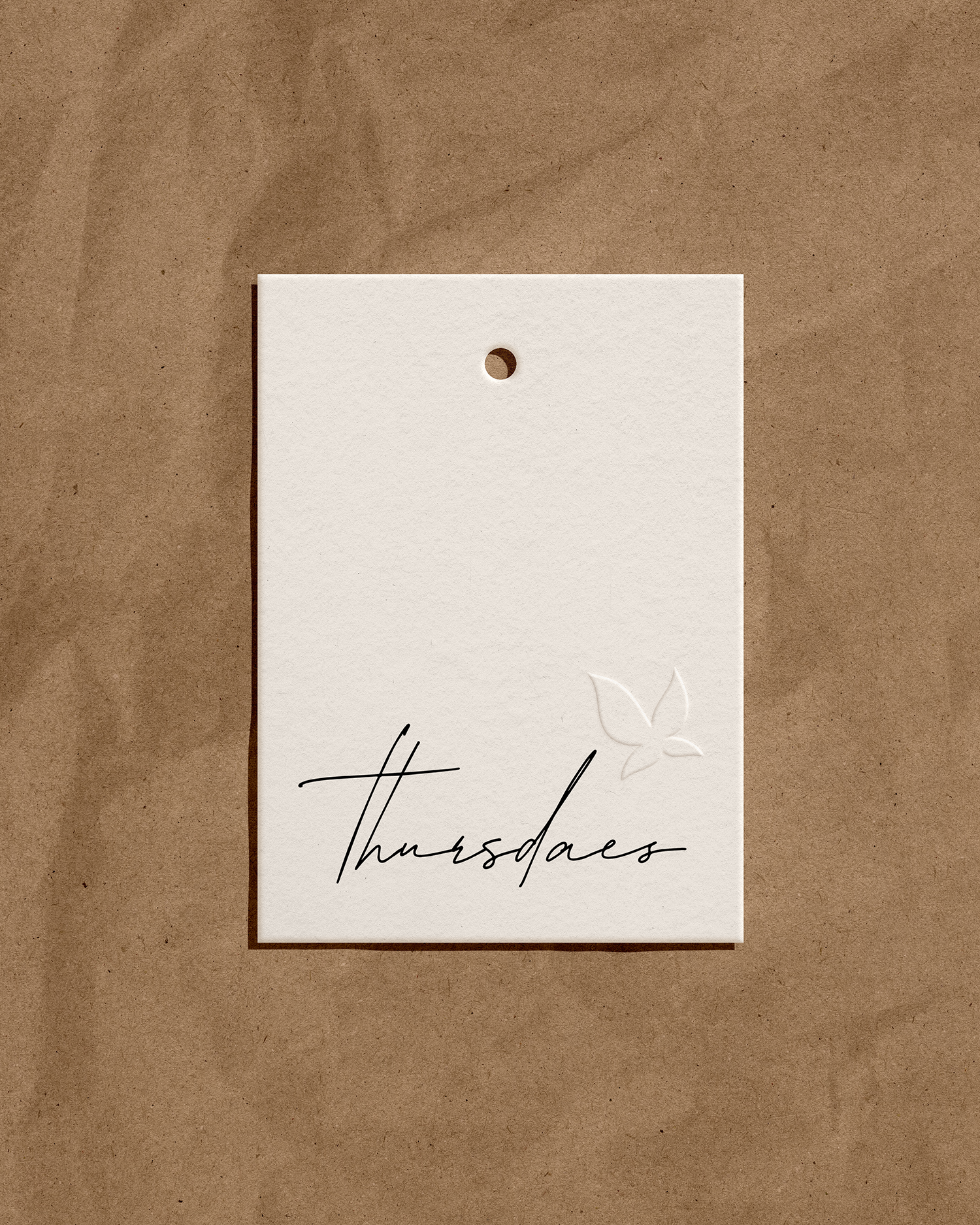 A white card with a small leaf outline and elegant handwritten text that reads 'Thursday'. The card is positioned on a brown textured surface with a small hole at the top center.