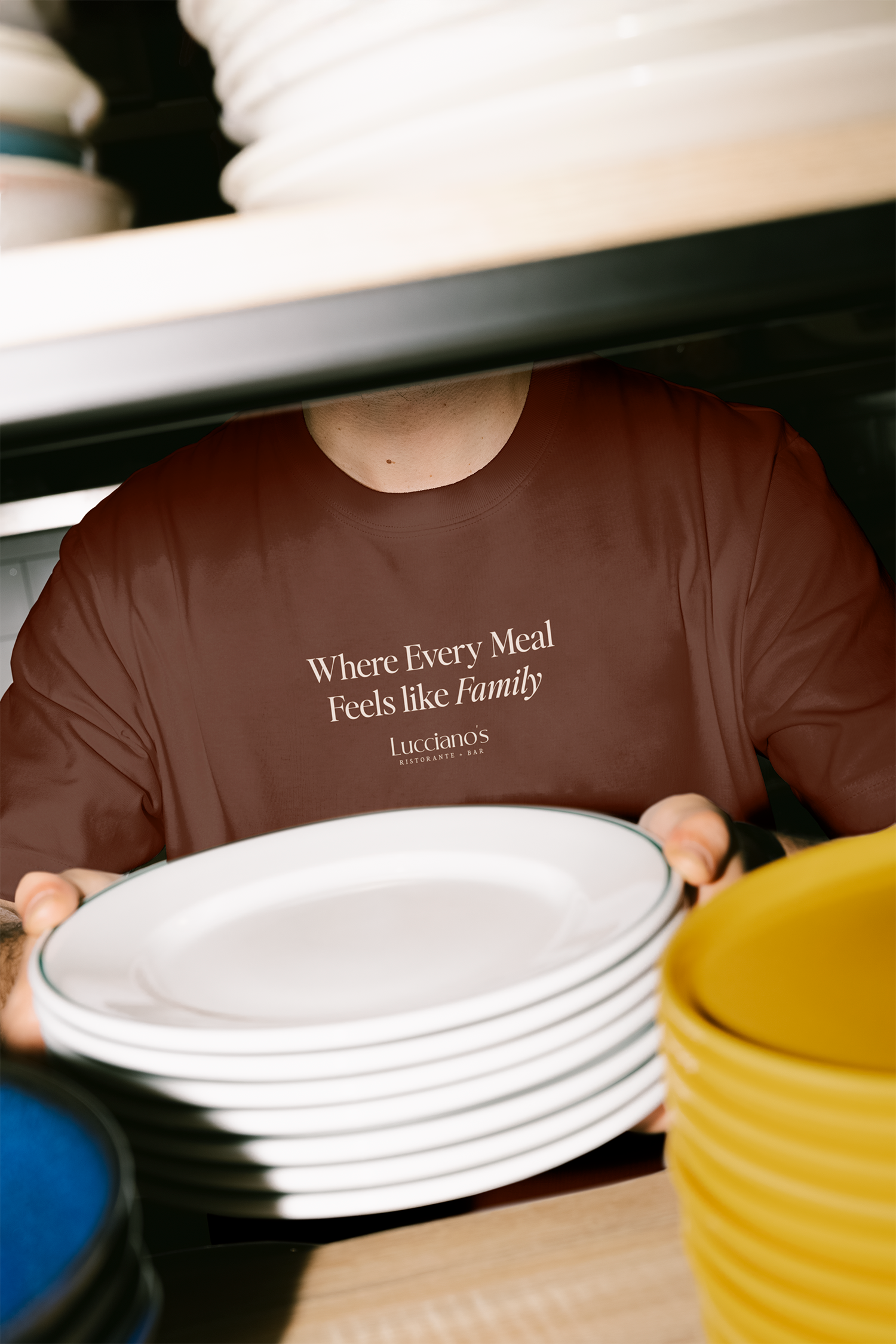 A person wearing a dark red T-shirt with the words 'Where Every Meal Feels like Family' printed on it, holding a white plate with a black rim, surrounded by stacks of white plates and bowls, with blue and yellow containers nearby.