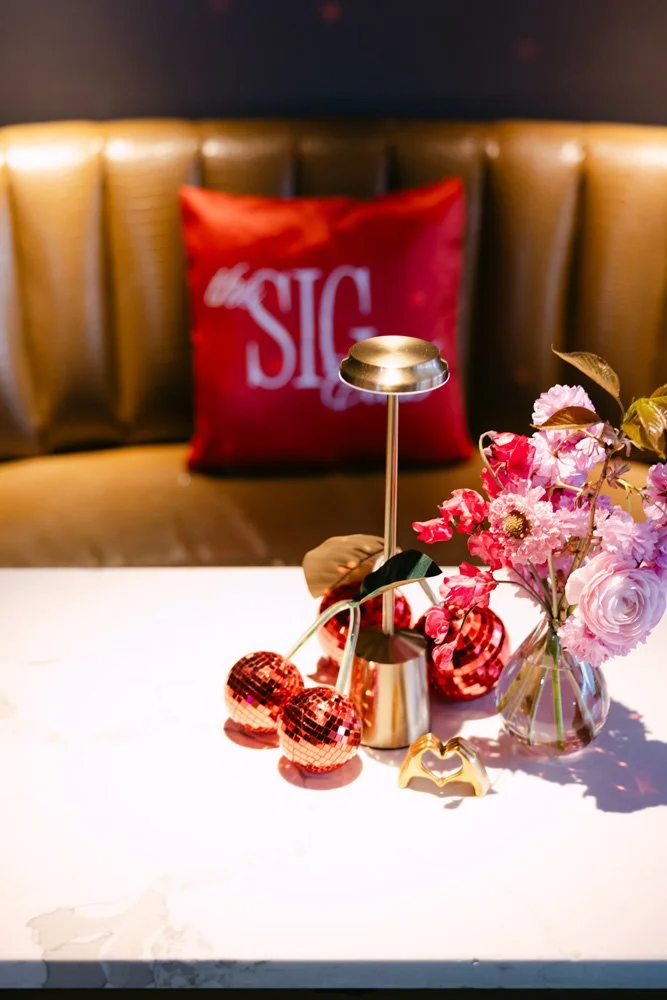 Table with a vase of pink and purple flowers, a small gold heart-shaped object, a lit modern lamp, and pink disco ball ornaments. In the background, a red pillow with white text is on a cushioned bench.