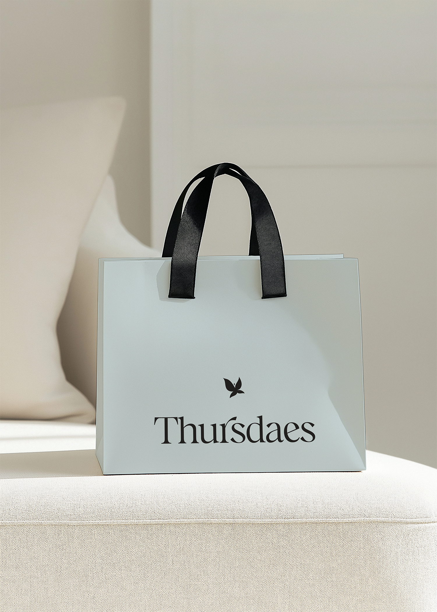A white shopping bag with black handles and the word 'Thursdays' with a leaf logo printed on it. The bag is on a neutral-colored surface with a light background.