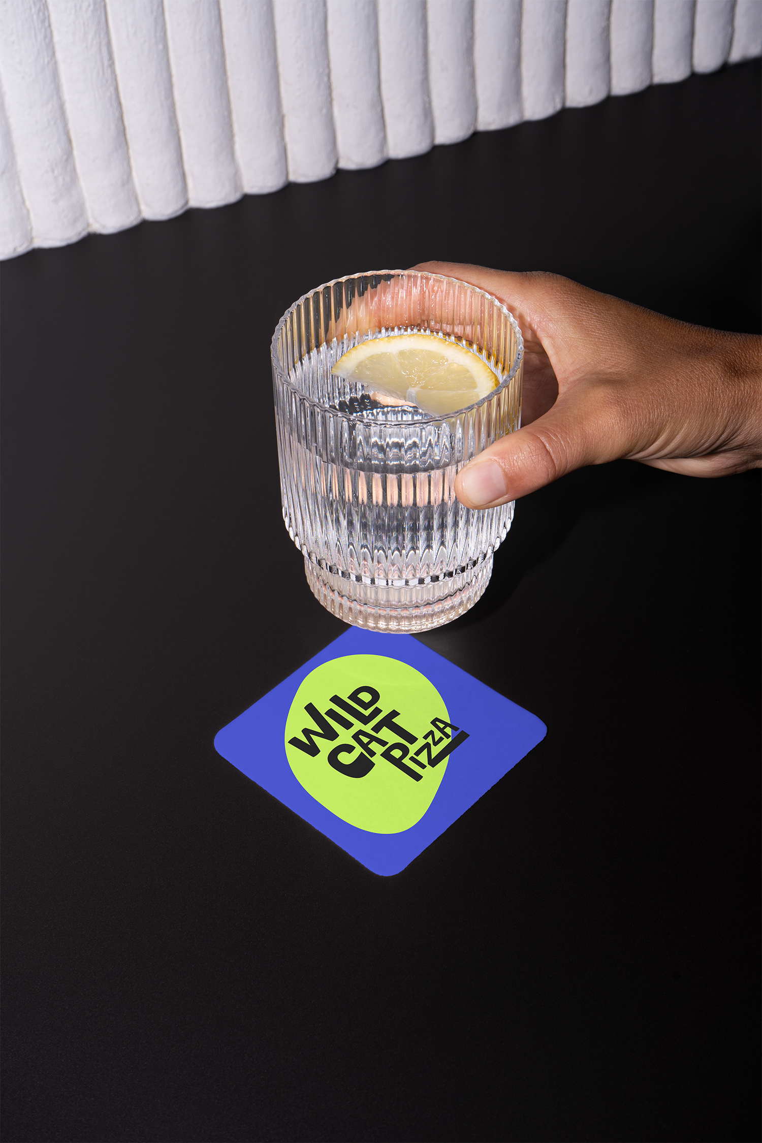 A clear glass of water with a lemon slice, held by a person's hand, resting on a black surface with a vibrant, colorful sticker that says 'WILD CAT PIZZA'.