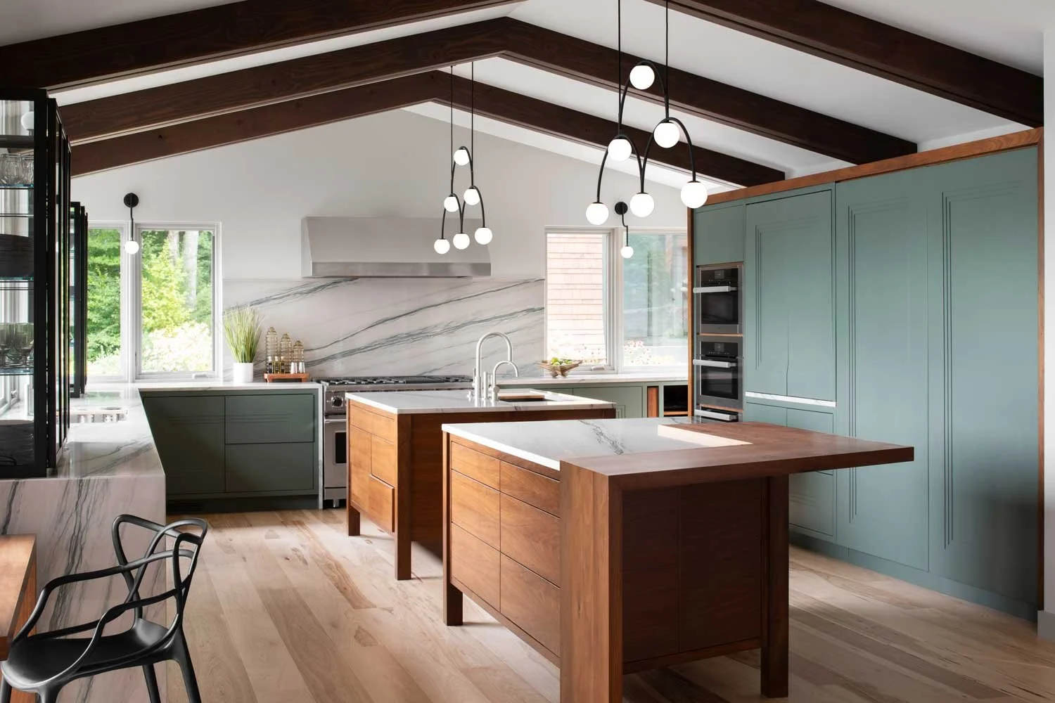 Modern kitchen with wooden beams, green and wooden cabinetry, island with white marble top, pendant lighting, and large windows showing greenery outside.