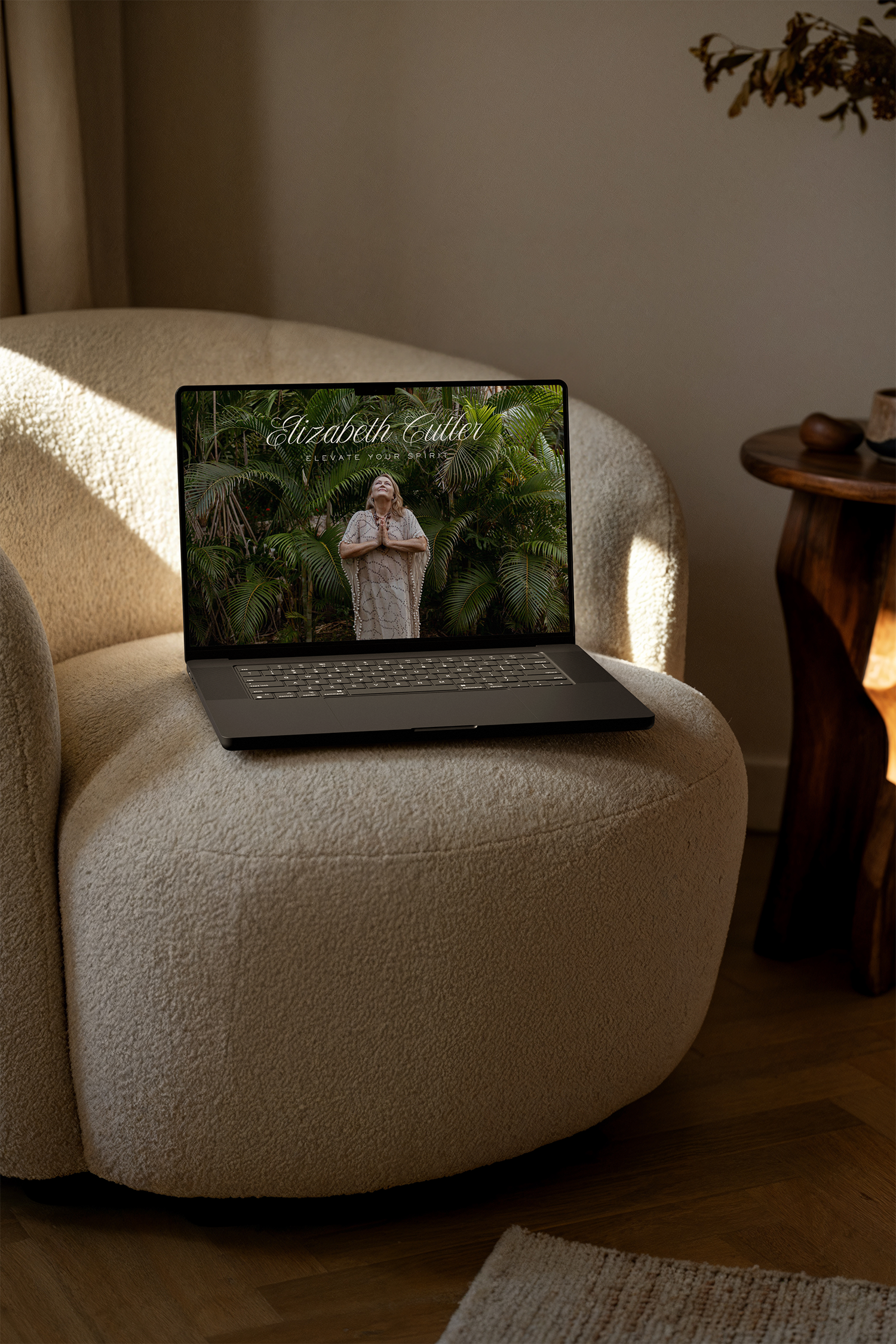 A beige armchair with a laptop on it displaying a woman in white surrounded by green plants and text that reads 'Elizabeth Cutler Elevate Your Spirit'. There is a small wooden side table with decorative objects next to the armchair, and a rug on a wo