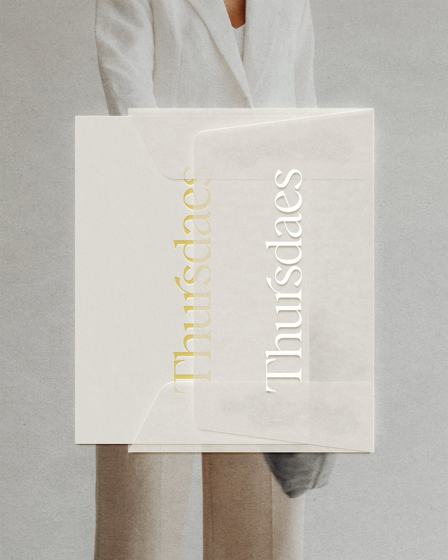 Close-up of a white envelope with cut-out gold and white text saying 'thru sadness' resting on a gray surface. Part of a person's legs and feet are visible underneath the envelope.