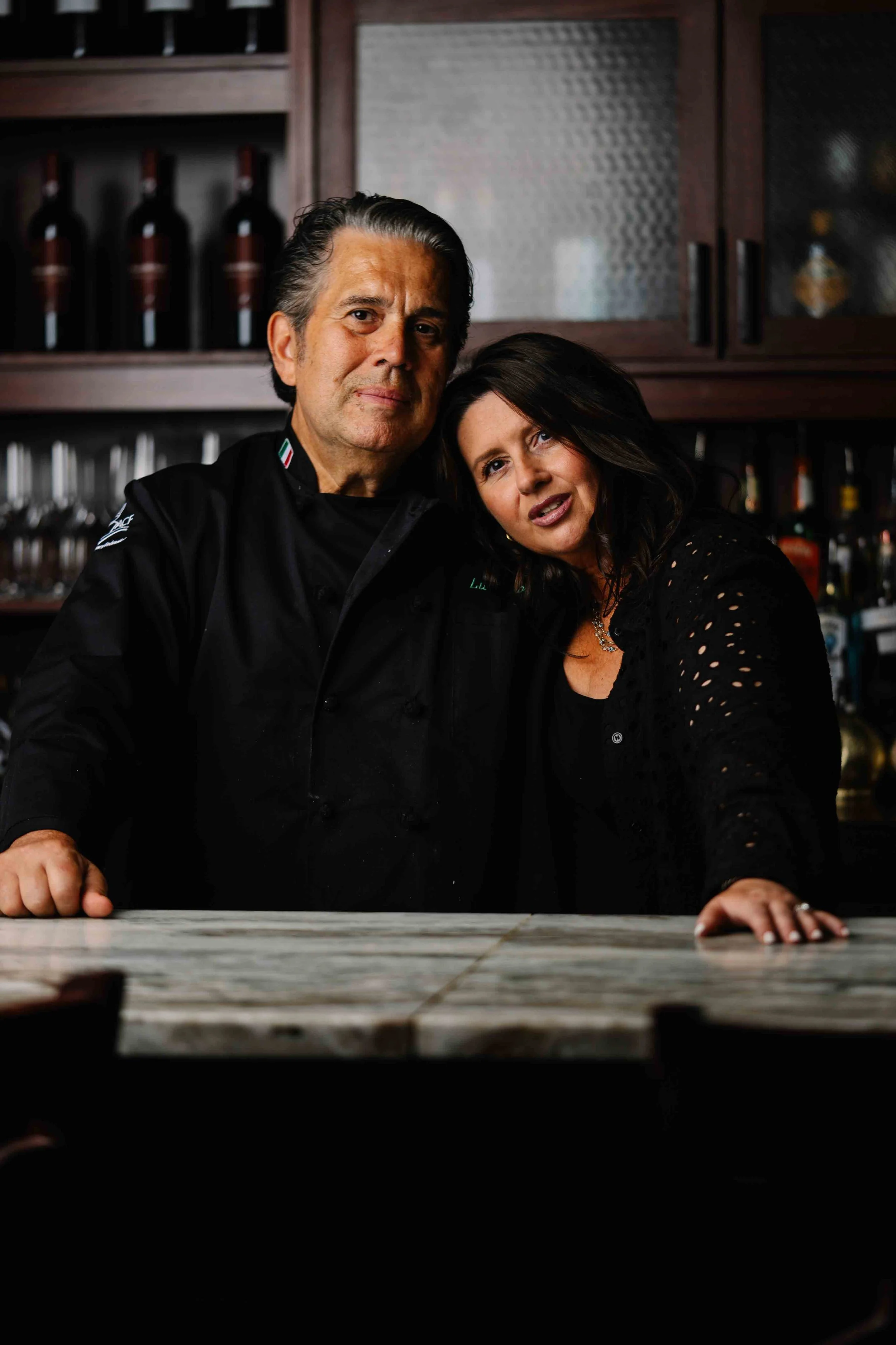 A man dressed as a chef and a woman are leaning against a wooden bar or counter with their hands resting on the surface. They are in a kitchen or bar setting with shelves of bottles in the background.