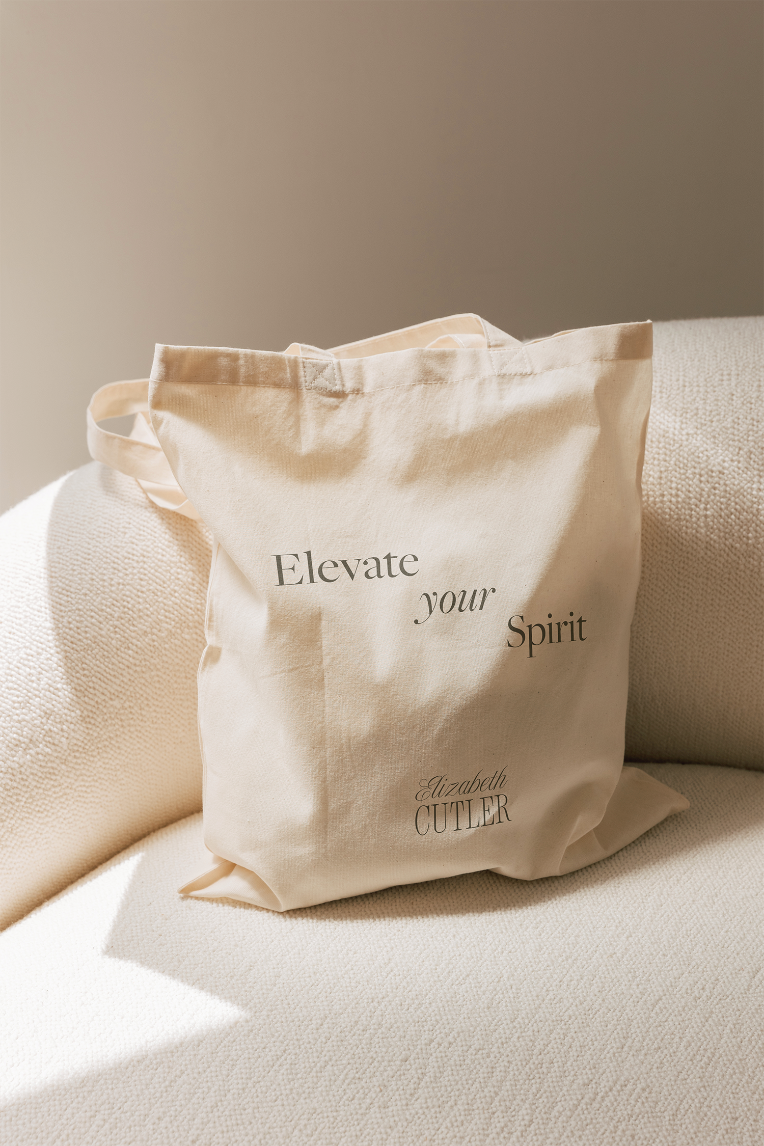 Cream-colored tote bag with the words 'Elevate your Spirit' and 'Elizabeth CUTLER' printed on it, resting on a beige textured armchair.