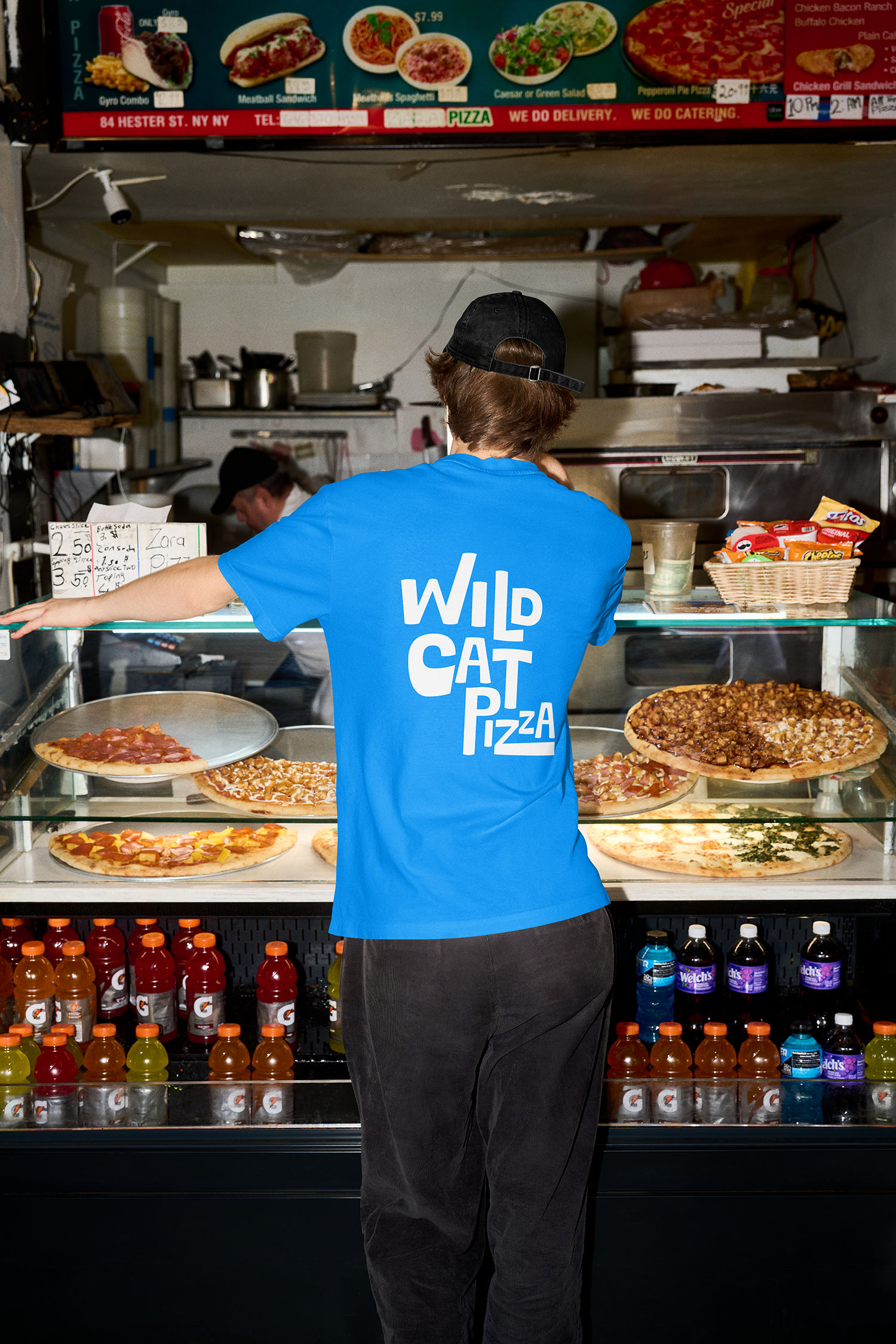 Person wearing a blue shirt and black cap standing in front of a pizza display case in a fast food restaurant. Several pizzas are visible inside, along with bottles of soda and snacks on shelves.